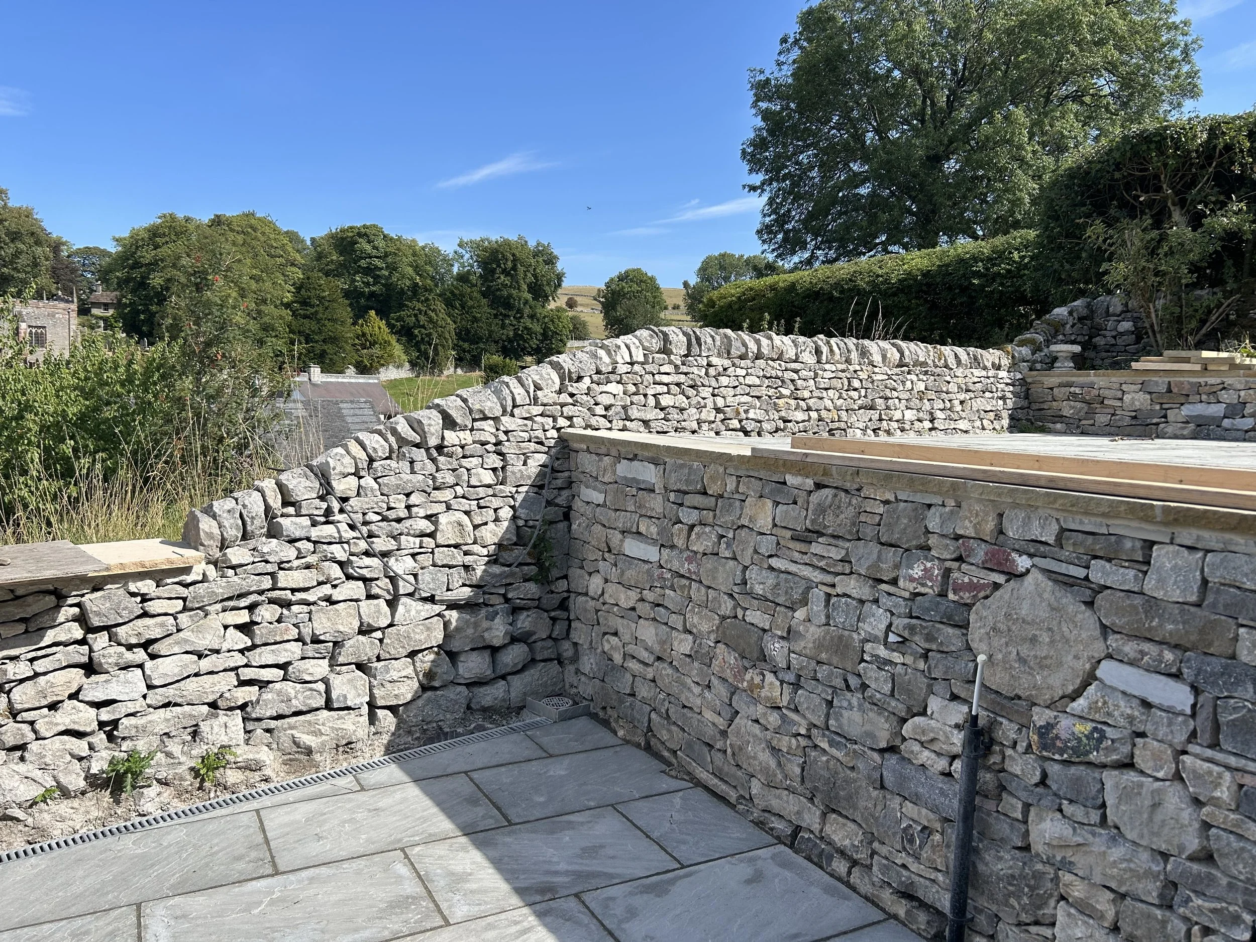 A newly built dry stone wall surrounds a patio area with light gray stone tiles, against a backdrop of green trees and a clear blue sky. Dry stone effect wall, gabion, breeze blocks, stick, mortar, stone , derbyshire