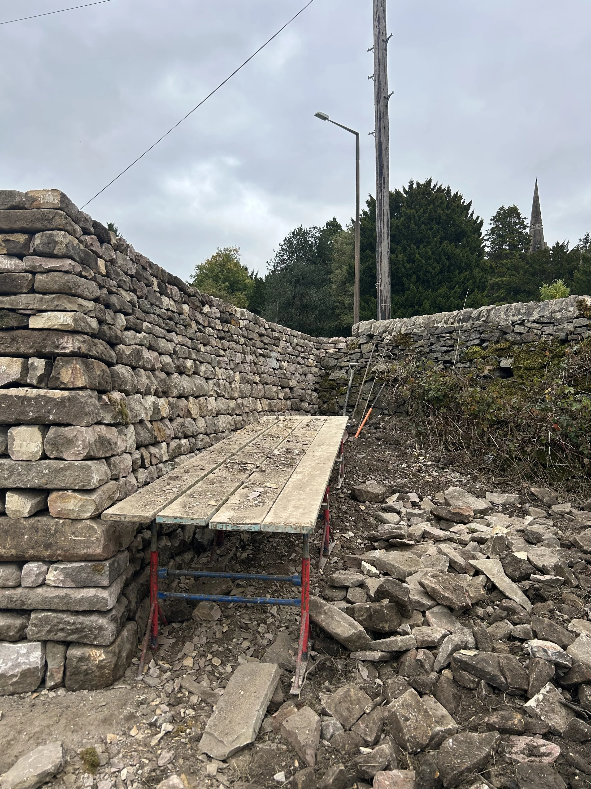 A stone retaining wall under construction with a wooden work platform in front, scattered stones on the ground, utility poles, a streetlight, trees, and a church steeple in the background under a cloudy sky. Tall wall, crossing joints, builder, const