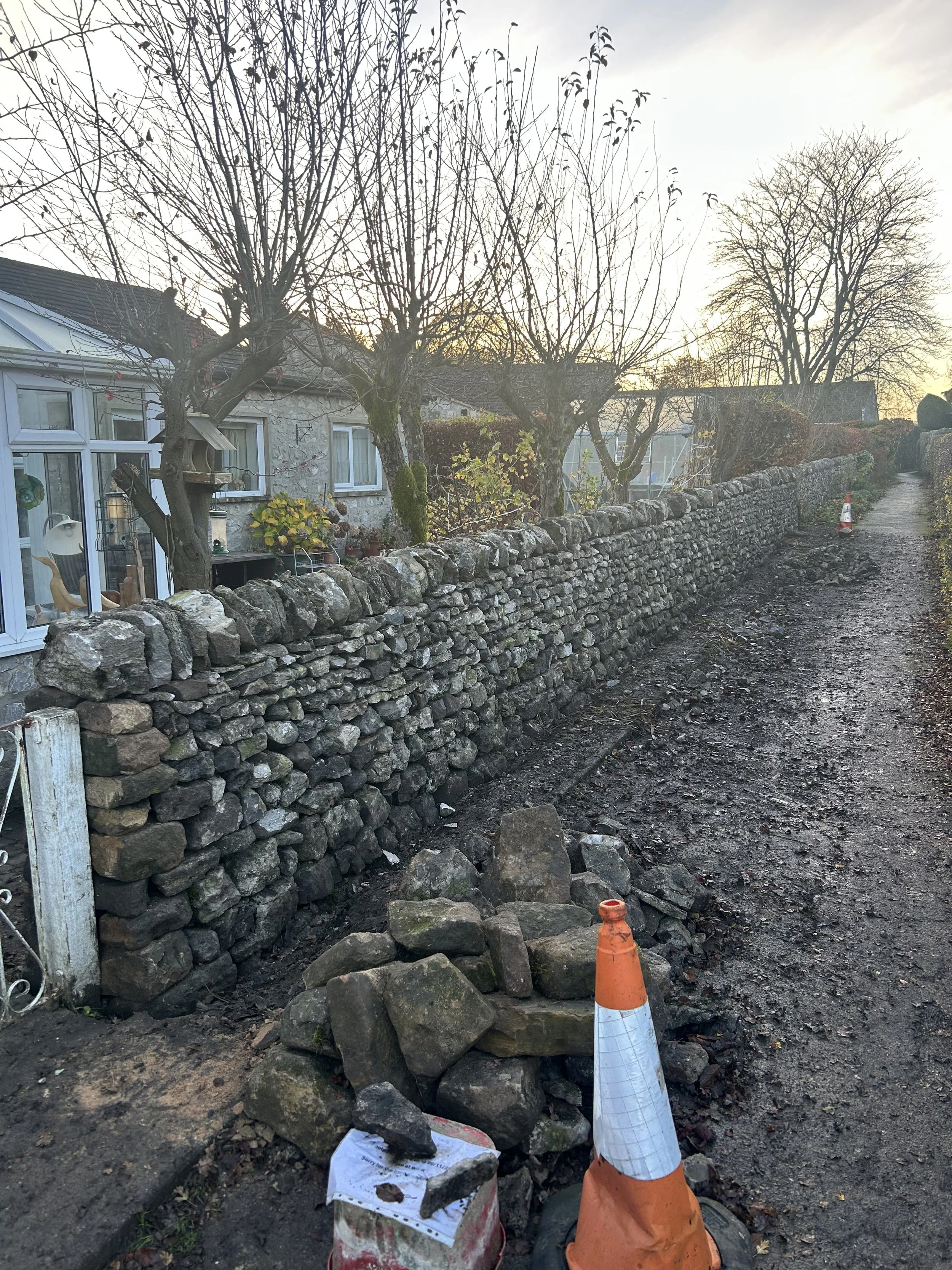 A DRY stone wall under construction runs along a dirt pathway beside a house with leafless trees. An orange and white traffic cone and loose stones are in the foreground. The scene appears to be during late fall or winter.stonework, dry stone WALLER