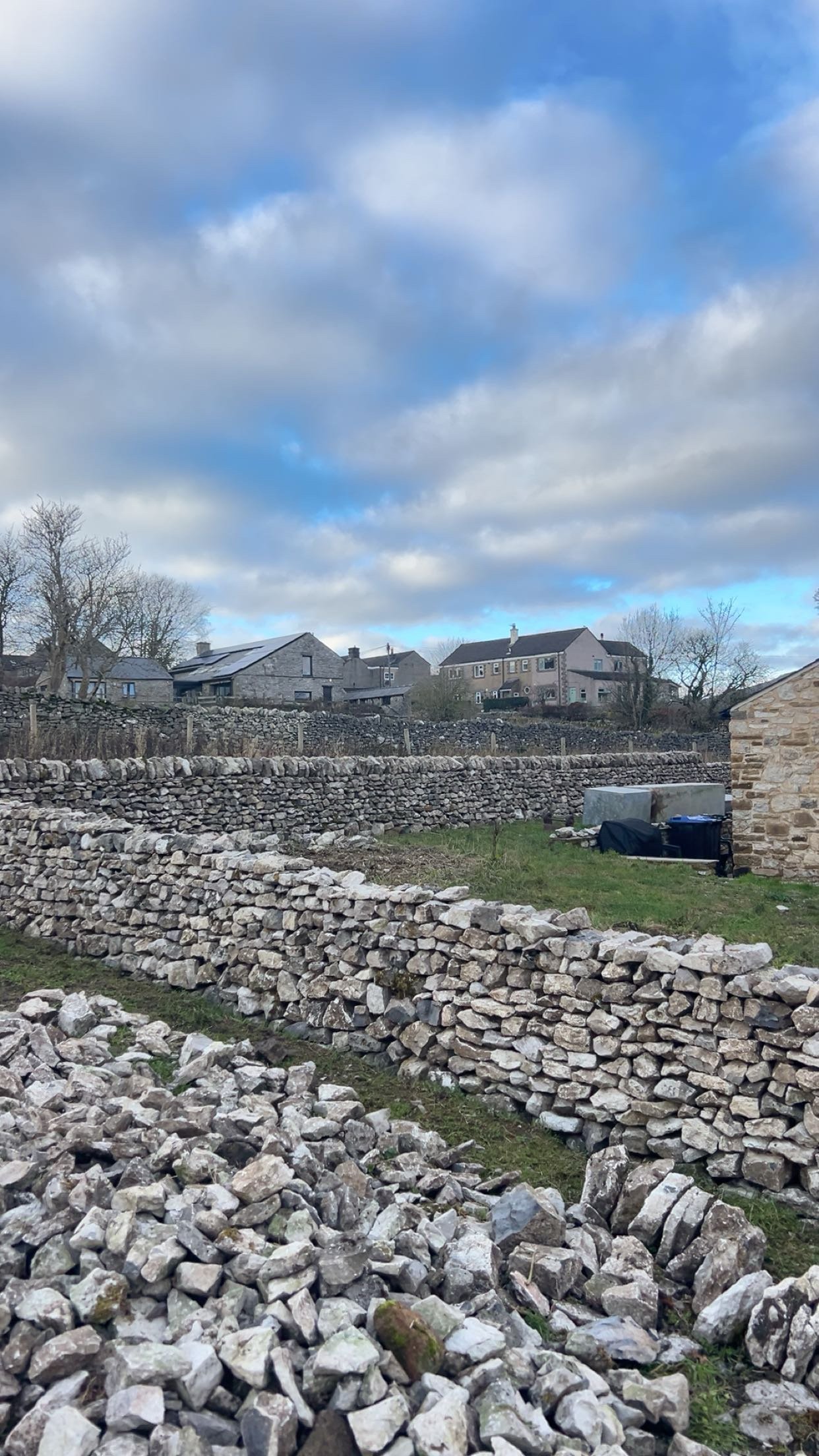 New build dry Stone wall terraces with stacked stones on a grassy area with houses in the background under a partly cloudy sky. New quarried stone, happy customers, bakewell, matlock, chesterfield, belper, ashbourne, derbyshire, uk, england
