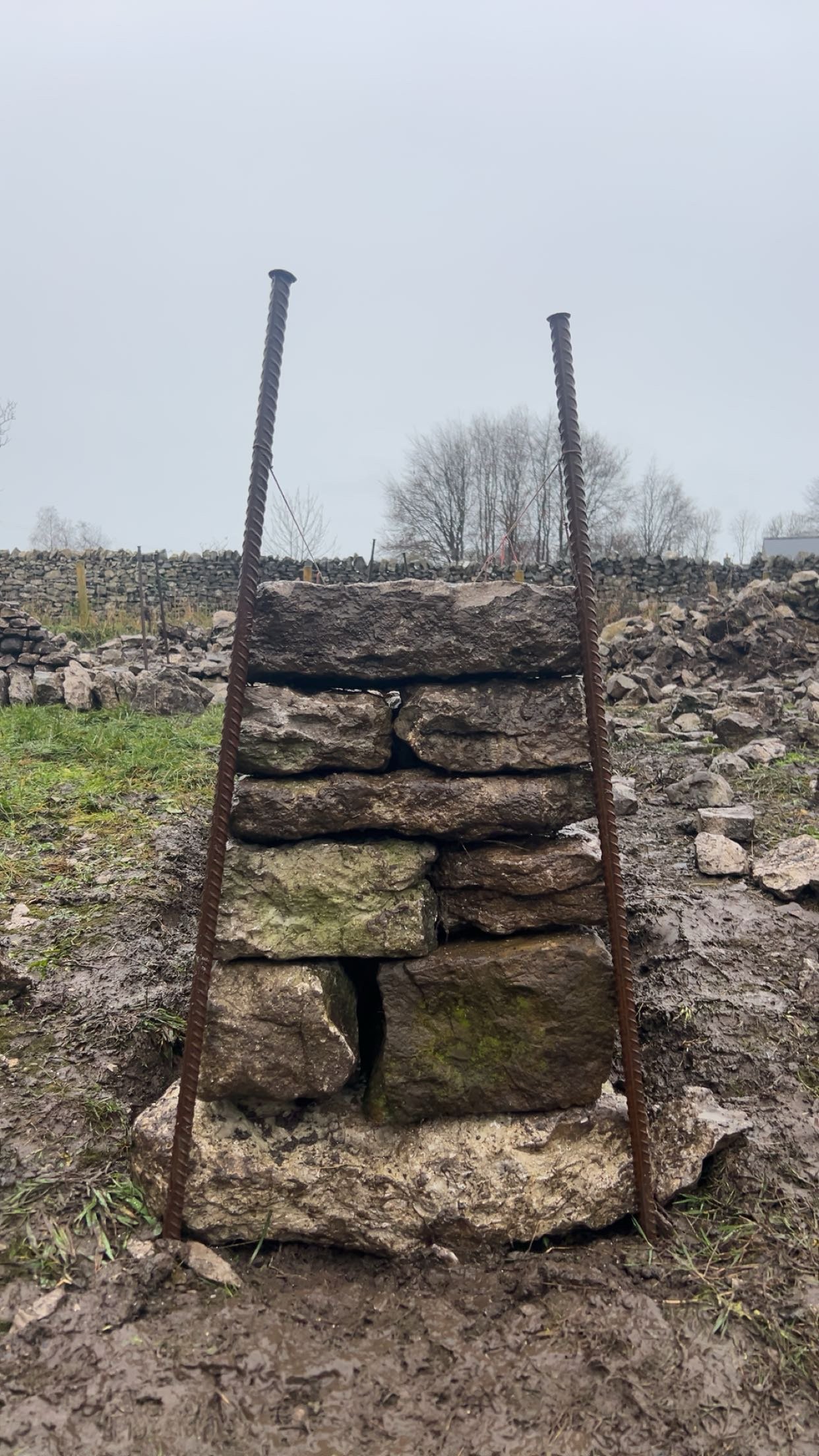 Stacked stones secured with rebar on muddy ground in an outdoor rocky landscape. Grant work, farming, cheekend, features, arch, pillars, dry stone pillars, drystone wall