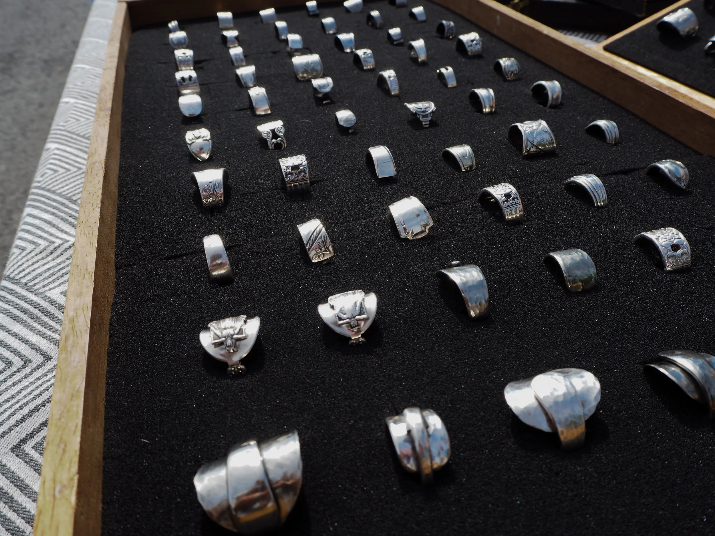 Various silver rings with engraved and decorative designs displayed on a black foam tray, some with ornate patterns and others with smooth surfaces.
