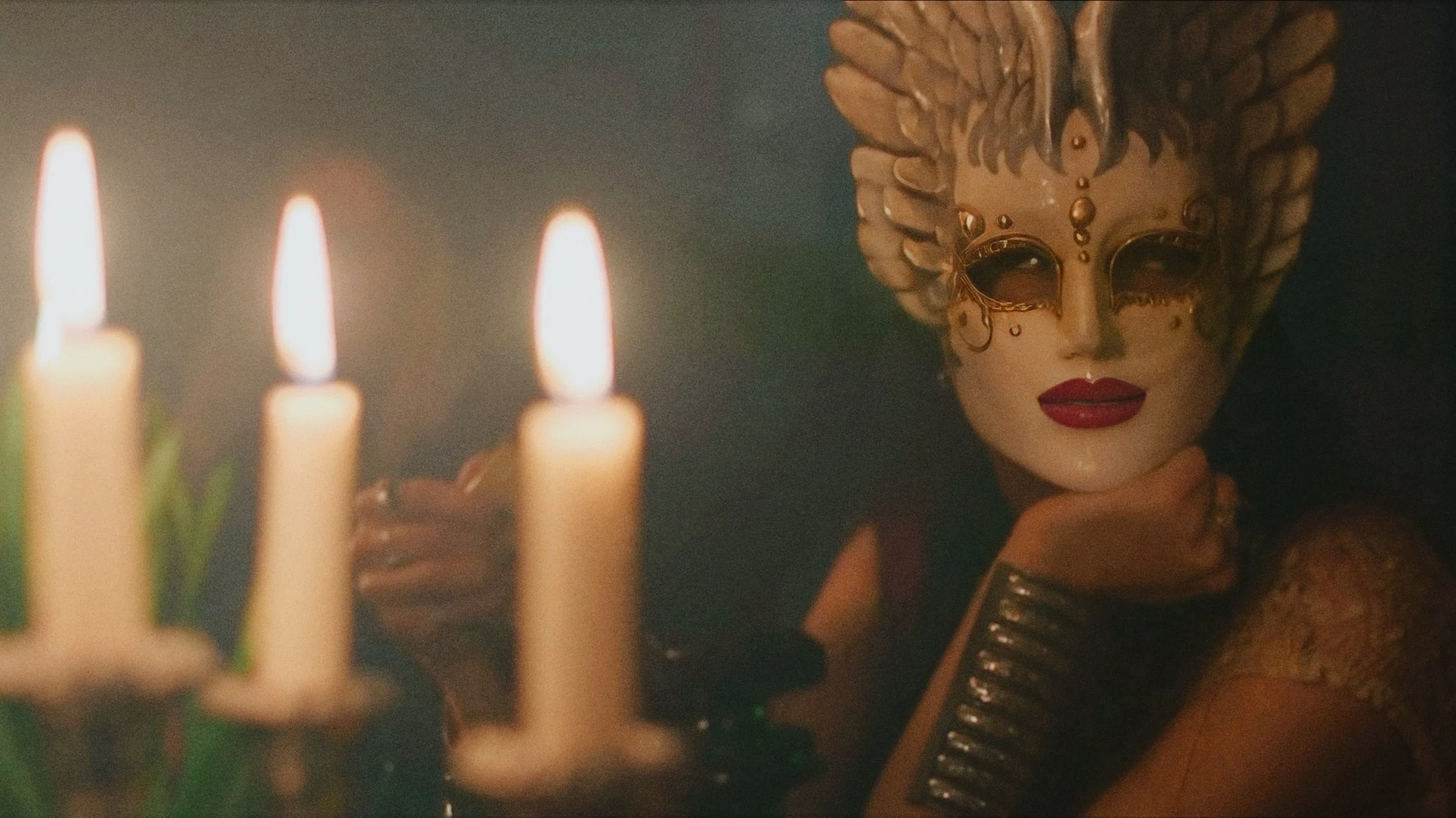 A person wearing an ornate, golden mask with wings on top, dark eye makeup, and red lipstick, looks directly at the camera while resting their chin on their hand. In the foreground, there are three lit candles. The scene is dark and atmospheric.