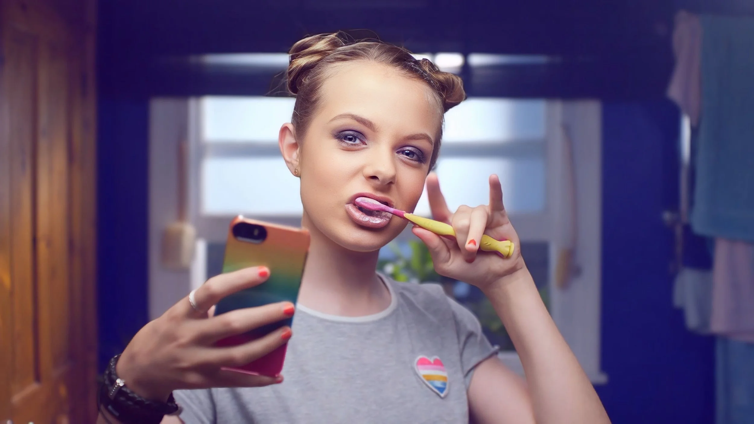Young woman with styled hair brushing her teeth and taking a selfie in a bathroom.