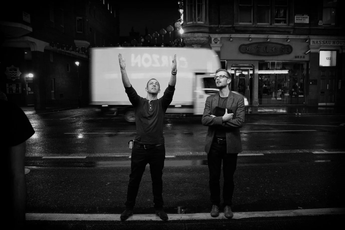 Two men standing on a wet city street at night, one with arms raised and looking up, the other with arms crossed, in front of a moving bus and illuminated shopfronts.
