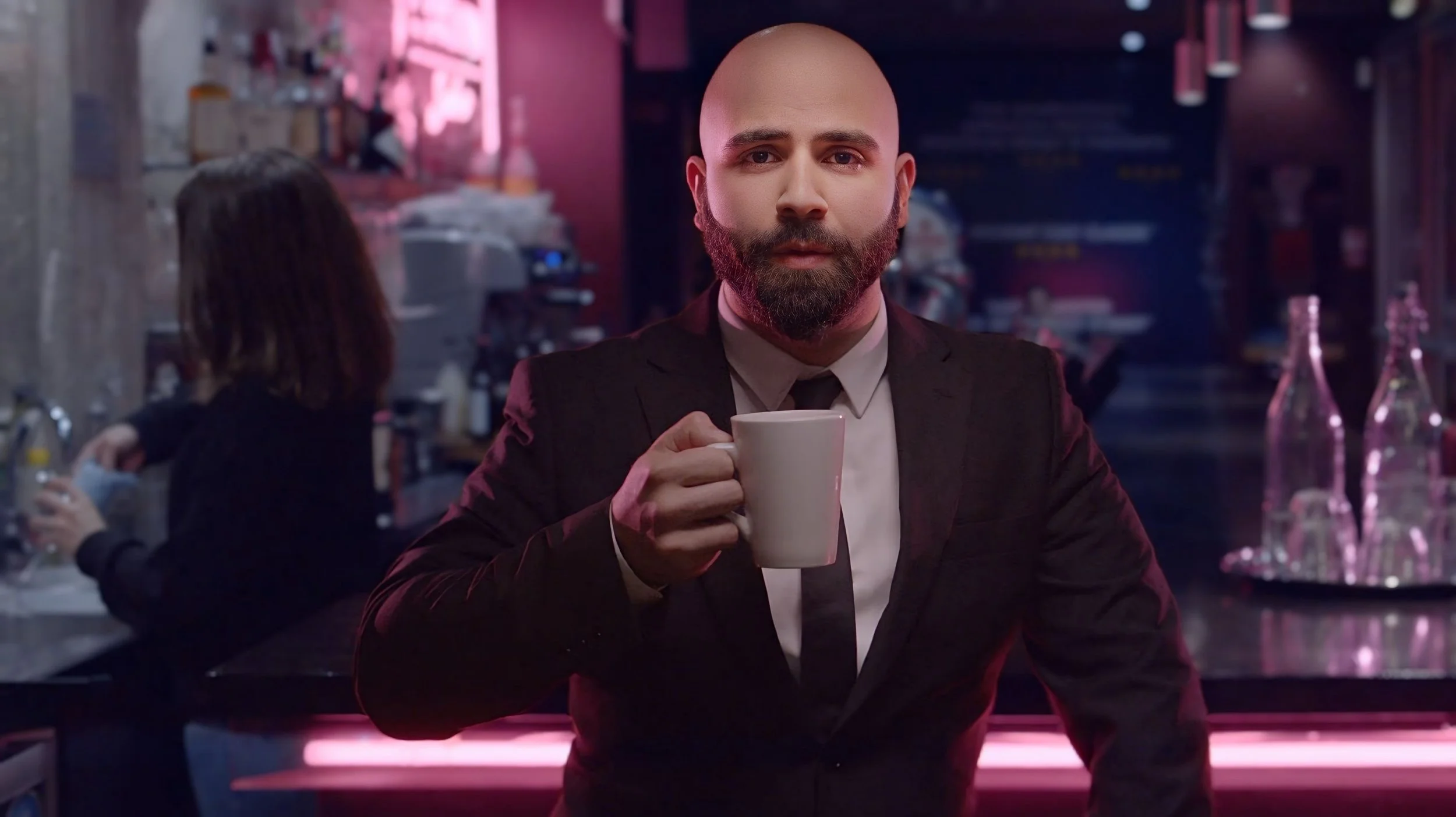 A man in a black suit and tie holding a white mug, standing in a dimly lit bar with pink lighting, with a woman in the background behind the bar.