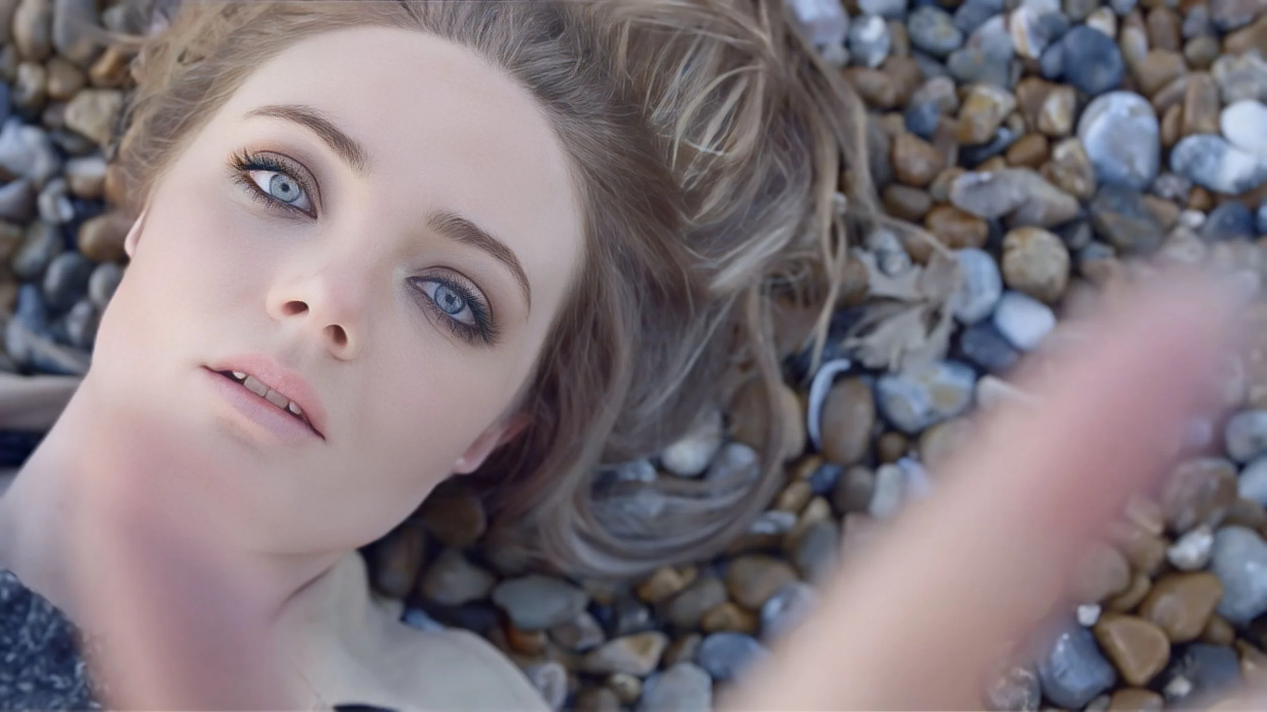 A young woman with blue eyes and light makeup lies on a bed of small, smooth stones, facing the camera.