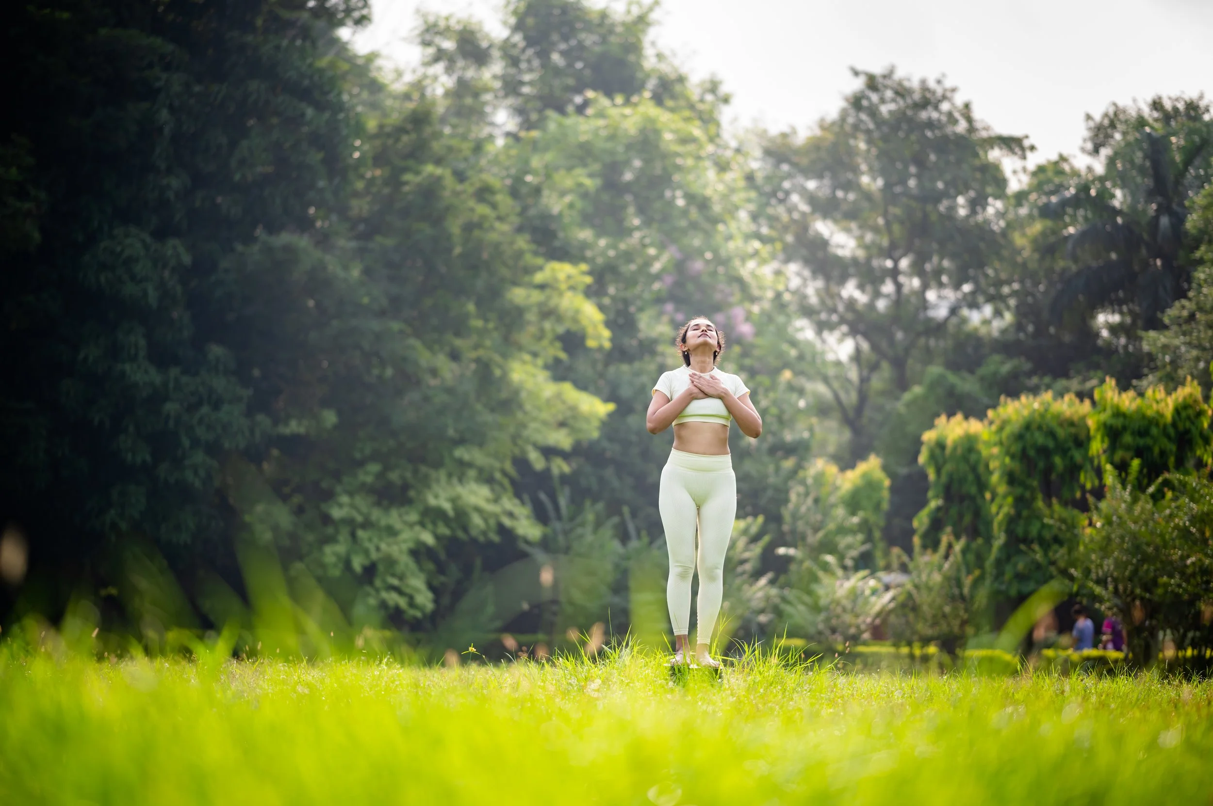 A woman wearing light yellow workout clothes standing on grass in a park, holding her hands to her chest and looking up with eyes closed, surrounded by green trees.