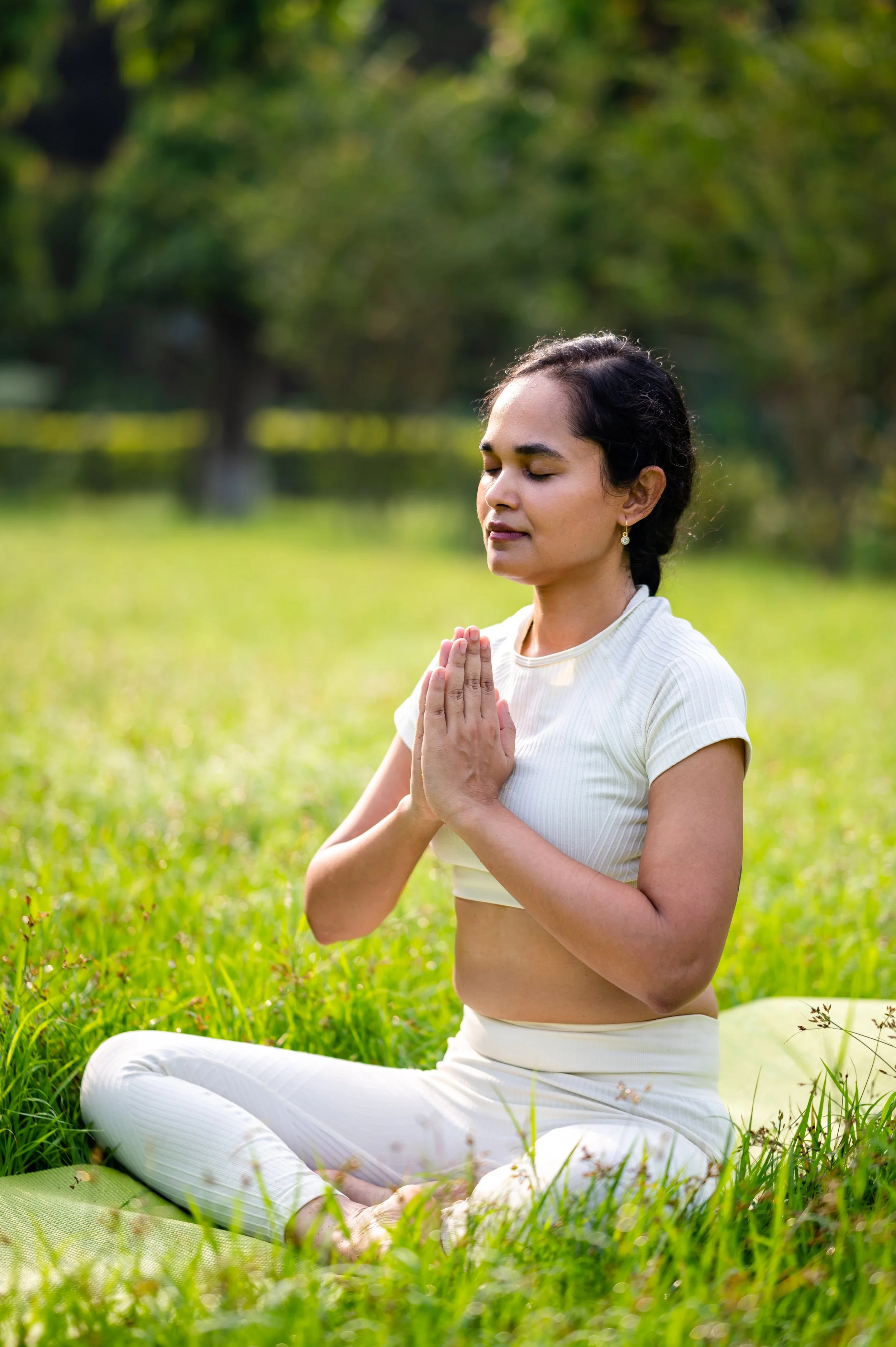 A woman practicing yoga outdoors in a grassy field, sitting cross-legged with hands in prayer position and eyes closed, enjoying a peaceful moment.