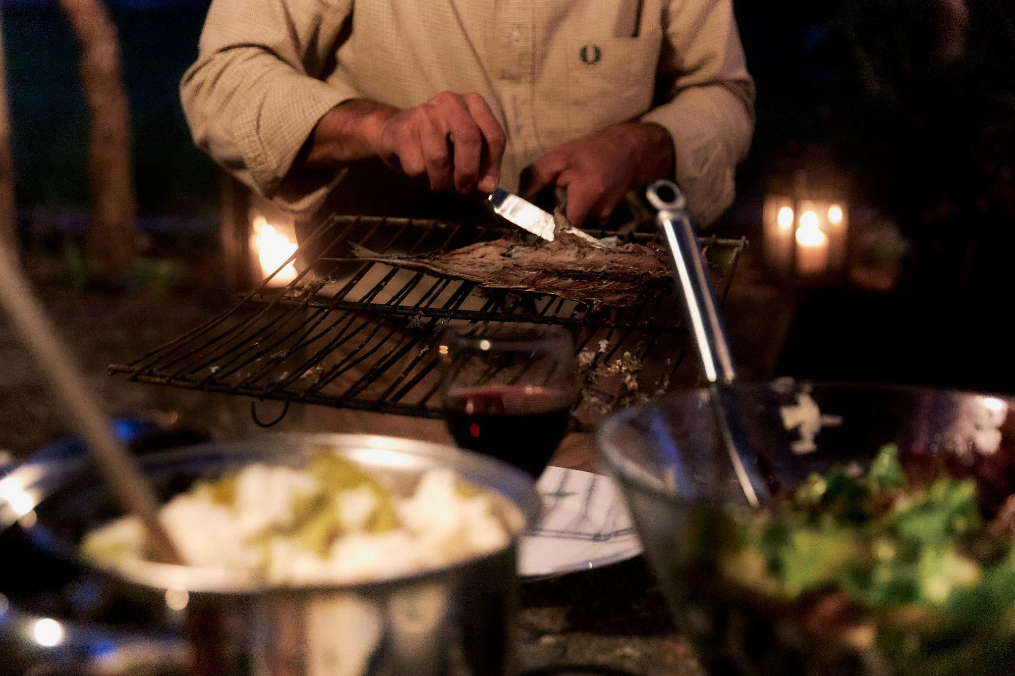 Person slicing cooked meat on a grill surrounded by food and drinks at night.