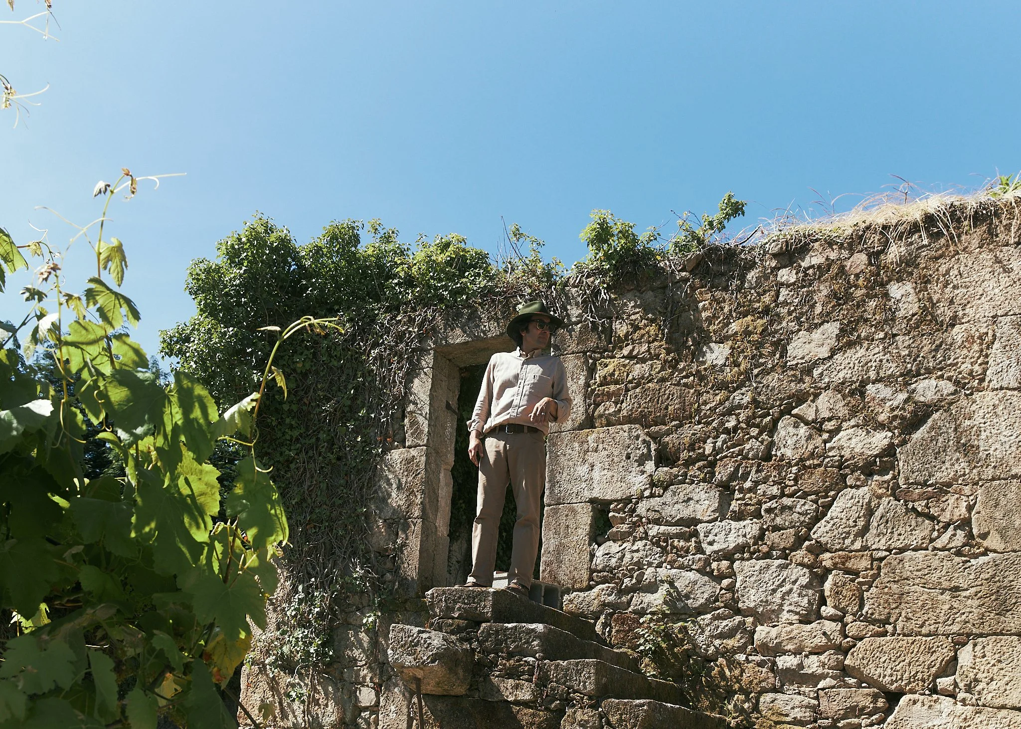 A woman wearing a tan outfit, hat, and sunglasses standing on stone steps outside ancient stone ruins against a clear blue sky.