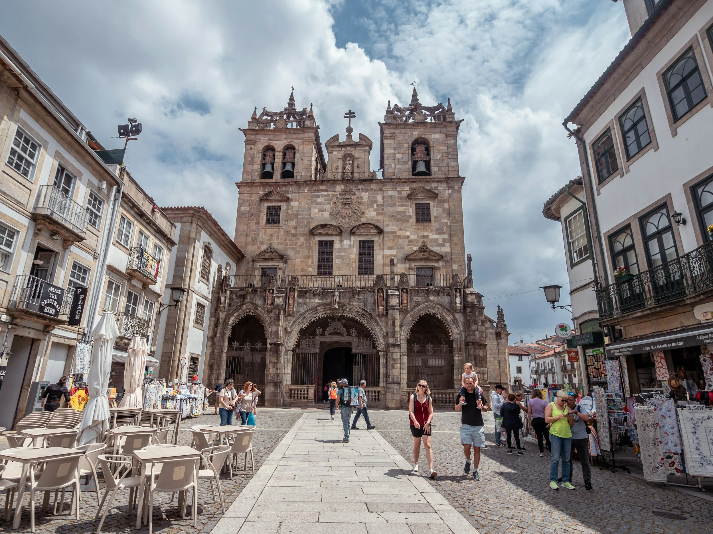 A historic stone church with twin towers in a busy cobblestone town square, surrounded by cafes, small shops, and pedestrians under a partly cloudy sky.