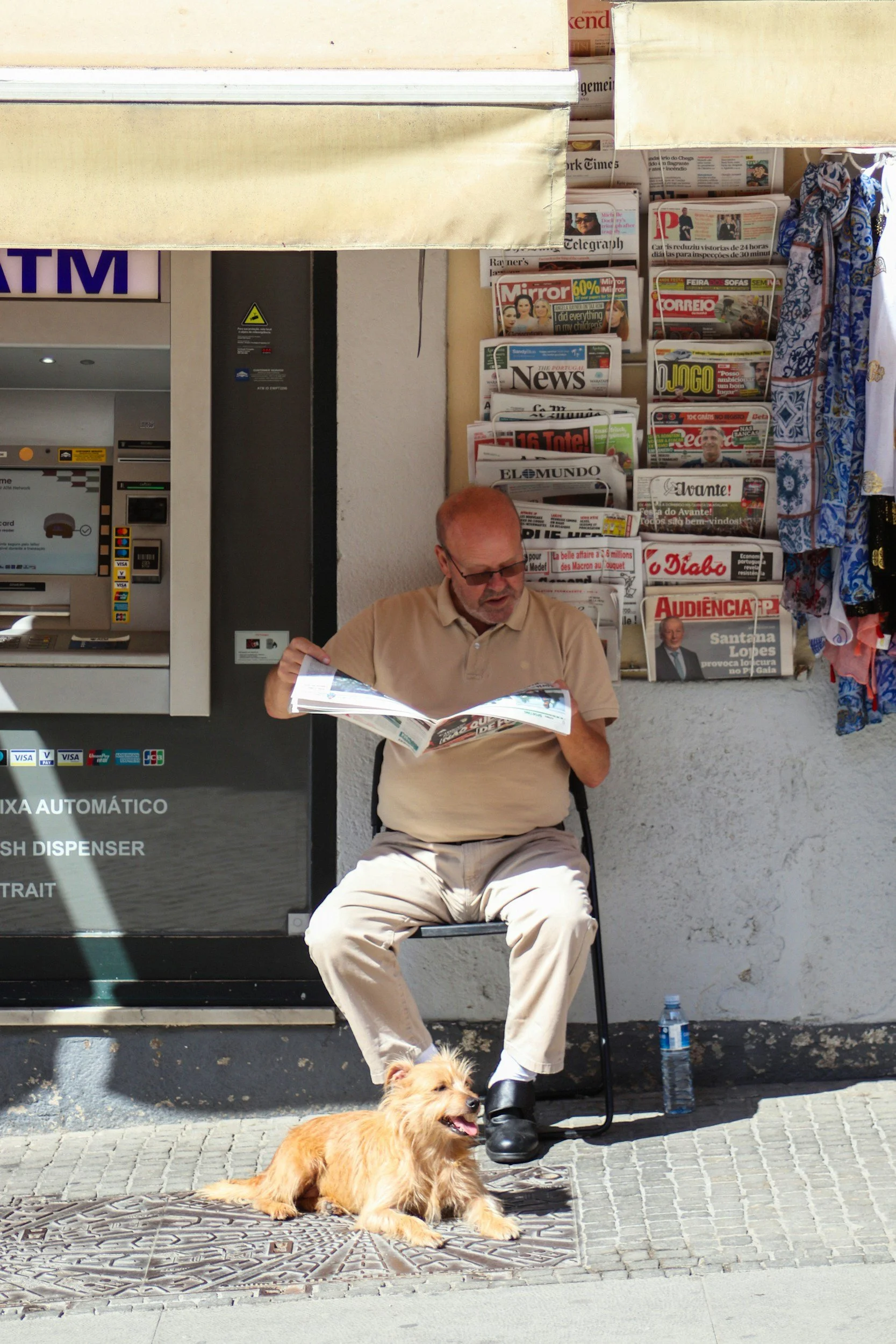 An elderly man with glasses reads a newspaper while sitting outside a small shop, with a small dog lying on the pavement nearby.