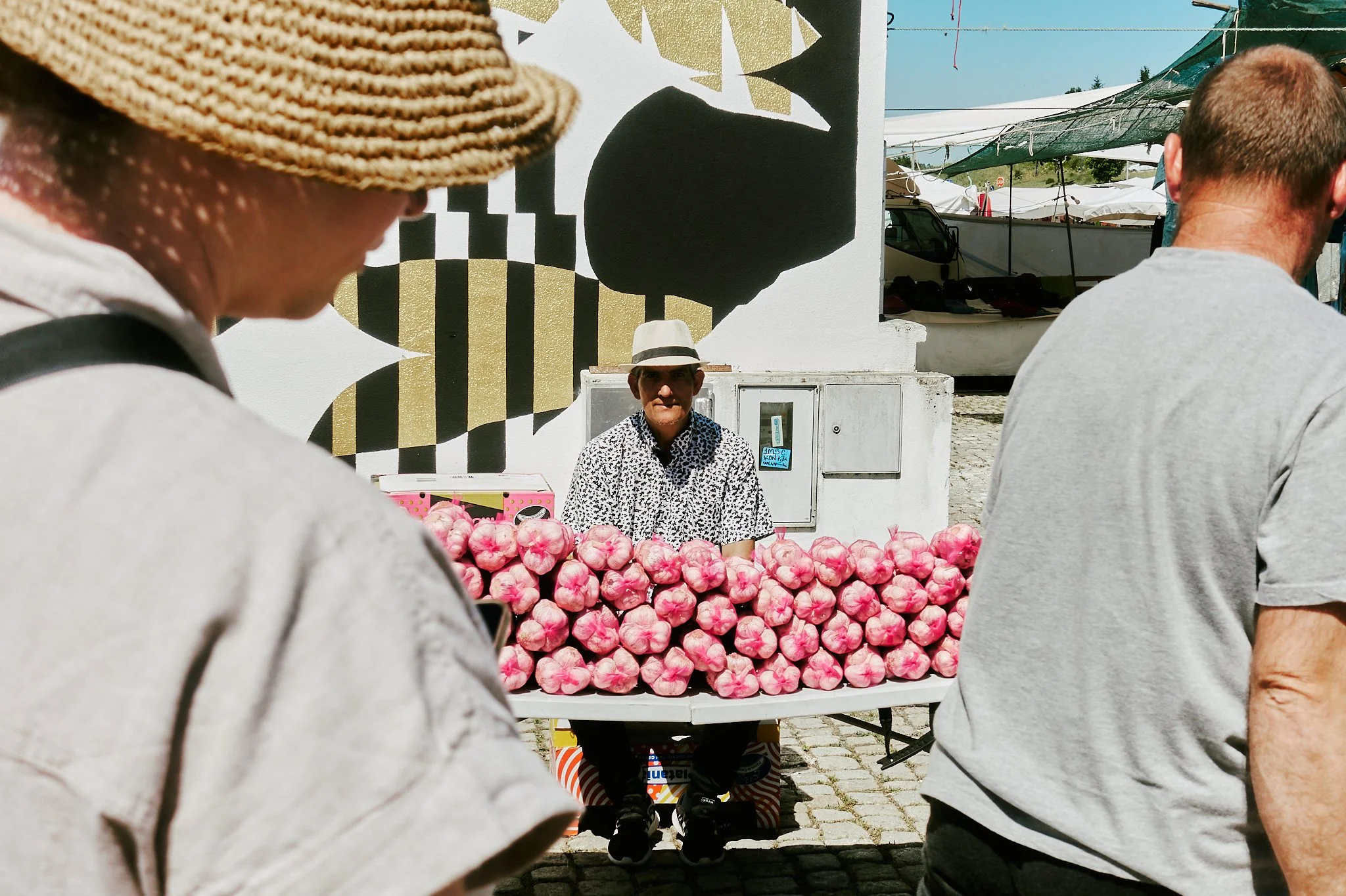 A street vendor sitting behind a table of pink garlic bulbs at an outdoor market, with tourists walking by and a mural on a wall behind him.