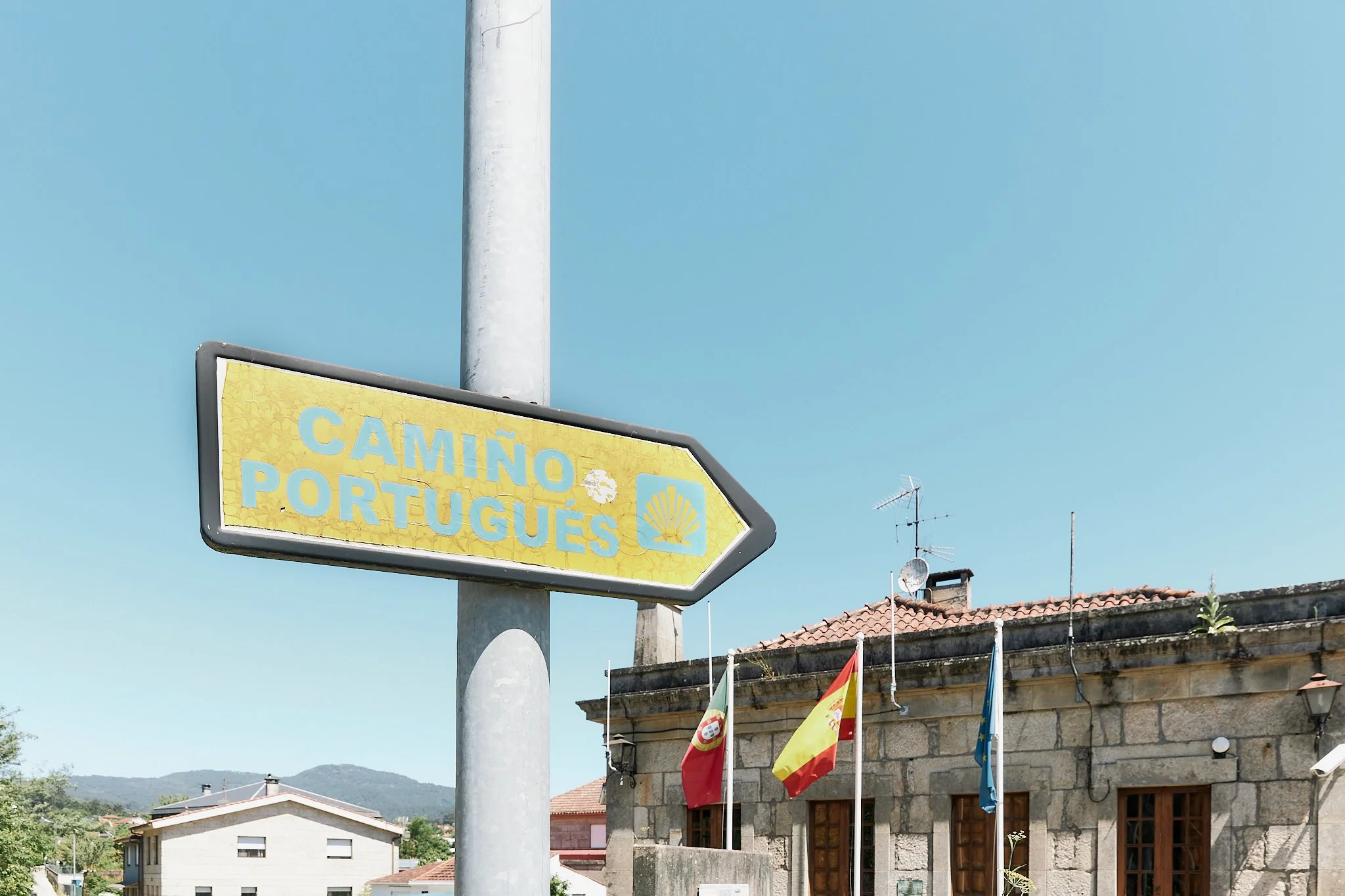 A yellow road sign with the words 'Camino Português' pointing to the right, mounted on a gray pole against a clear blue sky. In the background, there is a building with antennas and flags of Portugal, Spain, and the European Union.