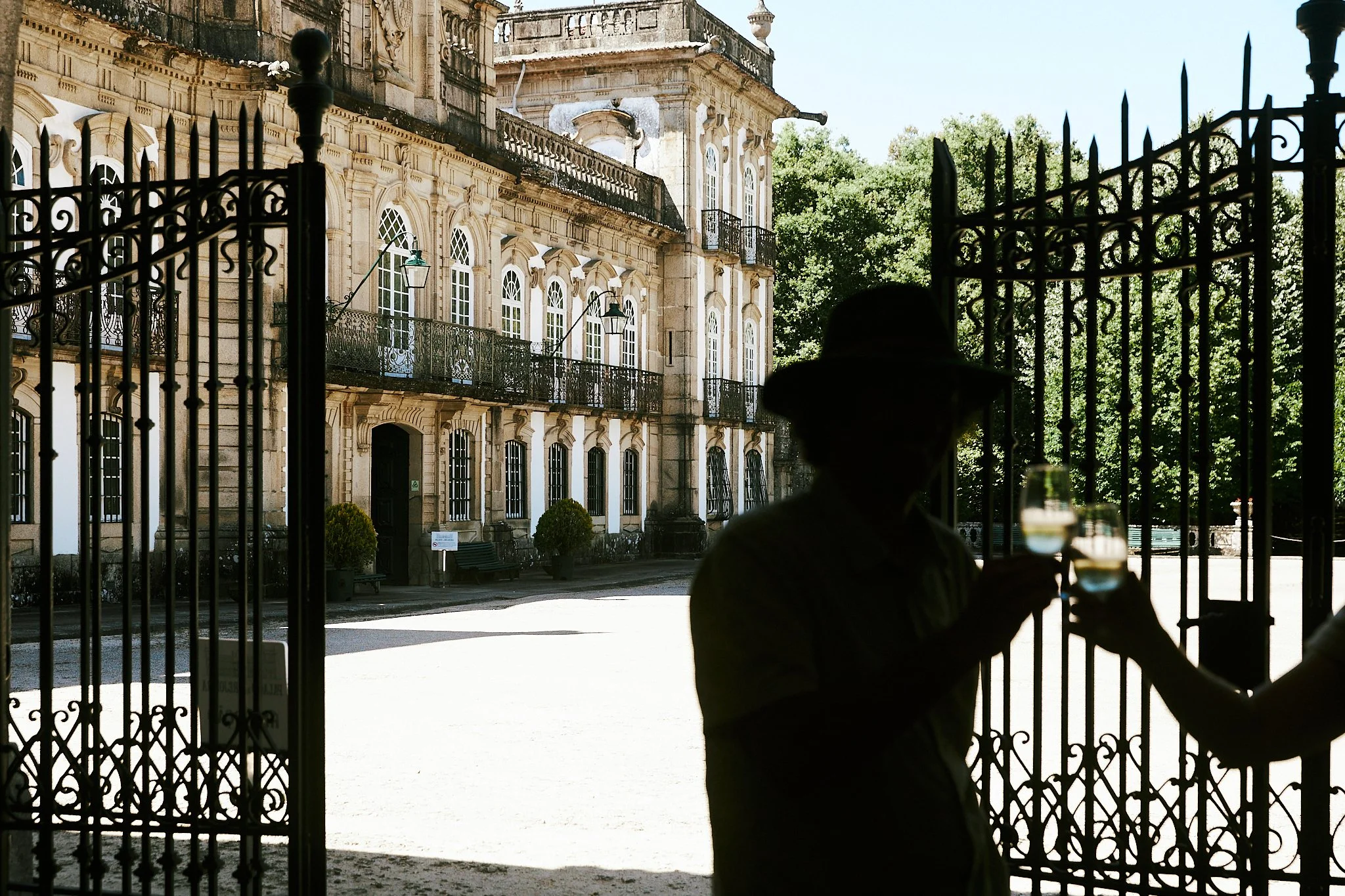 People enjoying wine behind a metal gate at a historic building with large windows and balconies, surrounded by lush green trees.