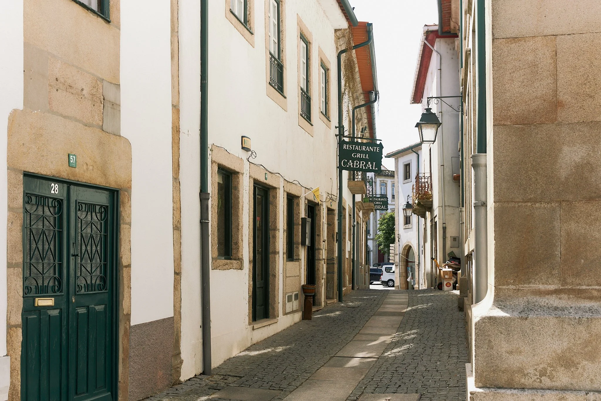 A narrow cobblestone street in a European town, lined with white and beige buildings with small windows and balconies. There are signs for a restaurant and grill called 'Cabral' hanging above the street.