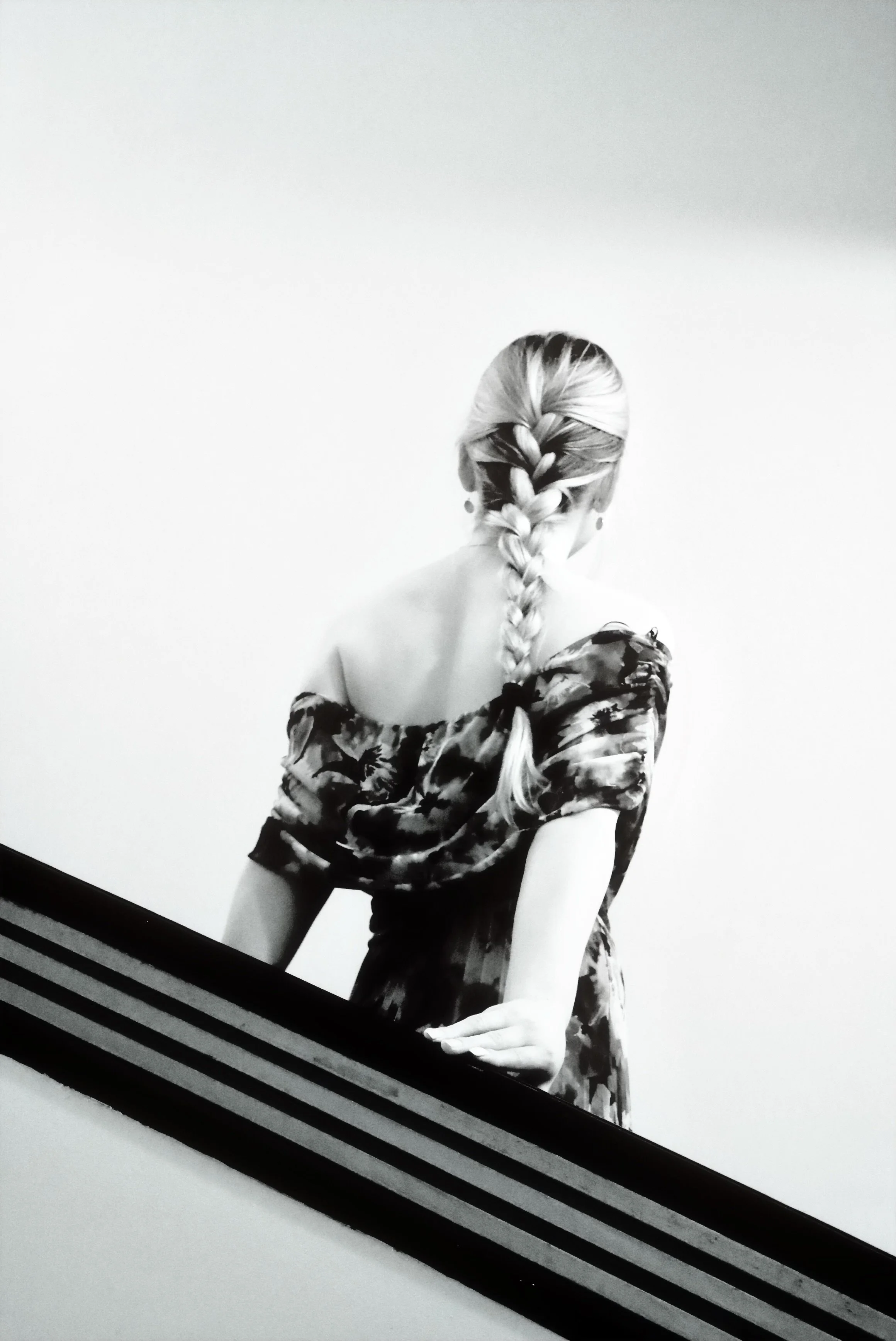 Back of a woman with a braided hairstyle, wearing a floral off-shoulder dress, leaning on a diagonal railing, against a plain light background.