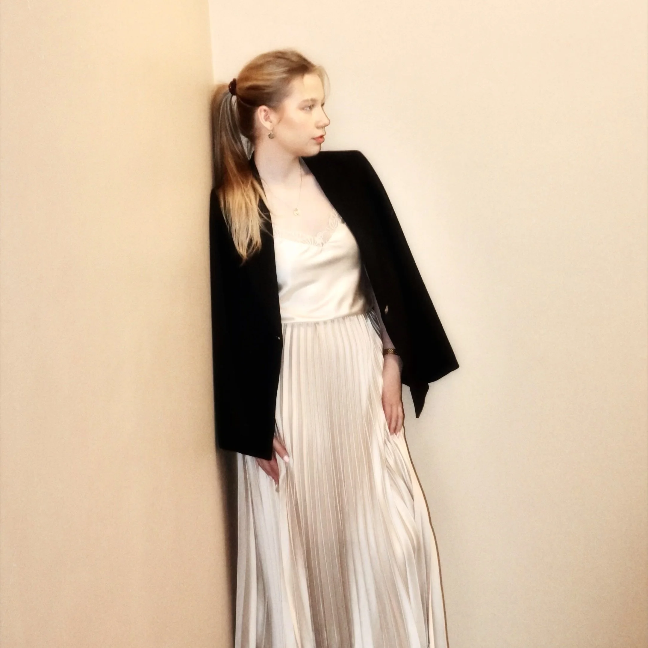 A young woman with light brown hair tied in a ponytail, wearing a black blazer over a white satin blouse and a long, pleated cream-colored skirt, leaning against a beige wall.