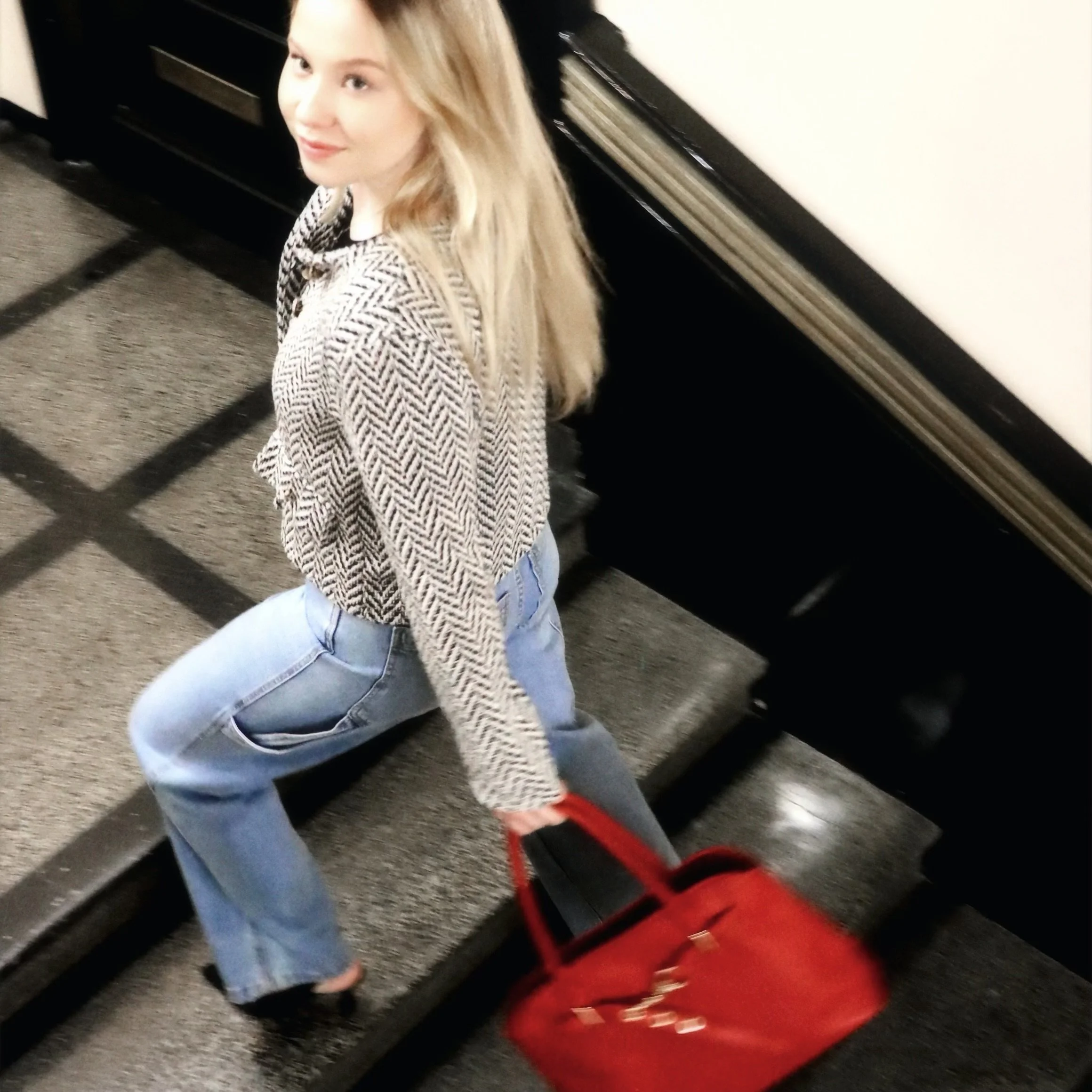 A woman with blonde hair in a patterned sweater and light blue jeans sitting on stairs, holding a red handbag.