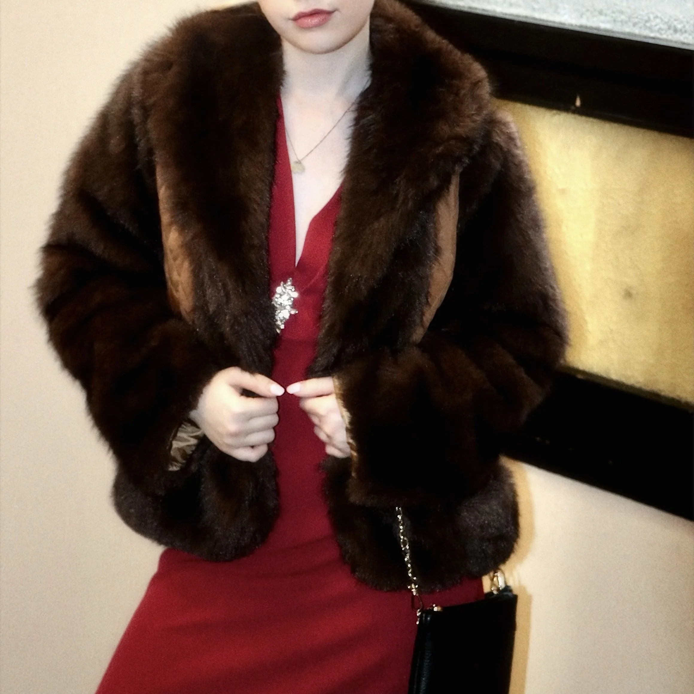 A woman wearing a red dress and a brown fur coat, holding the coat closed with her hands, with a black purse hanging from her shoulder.