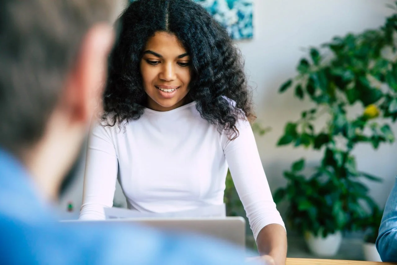 A woman with curly black hair smiling while looking at a laptop, with two blurred people in the foreground and a houseplant in the background.