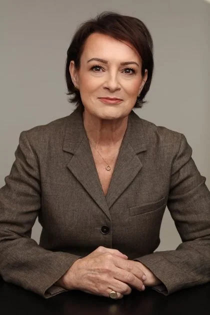 A woman with short dark hair and light skin, wearing a brown blazer, sitting at a table with her hands clasped.