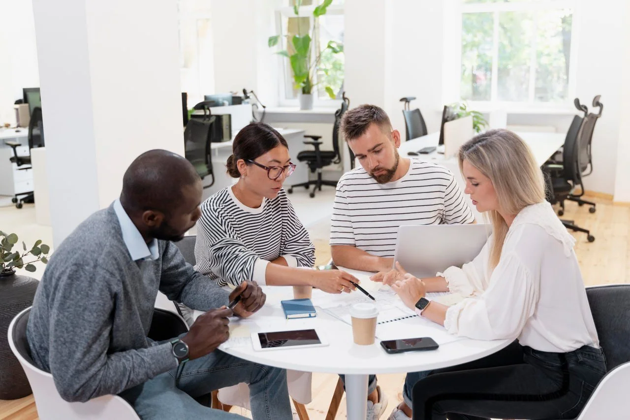 A diverse group of five people having a meeting around a white table in an office, with laptops, tablets, notebooks, and coffee cups on the table.