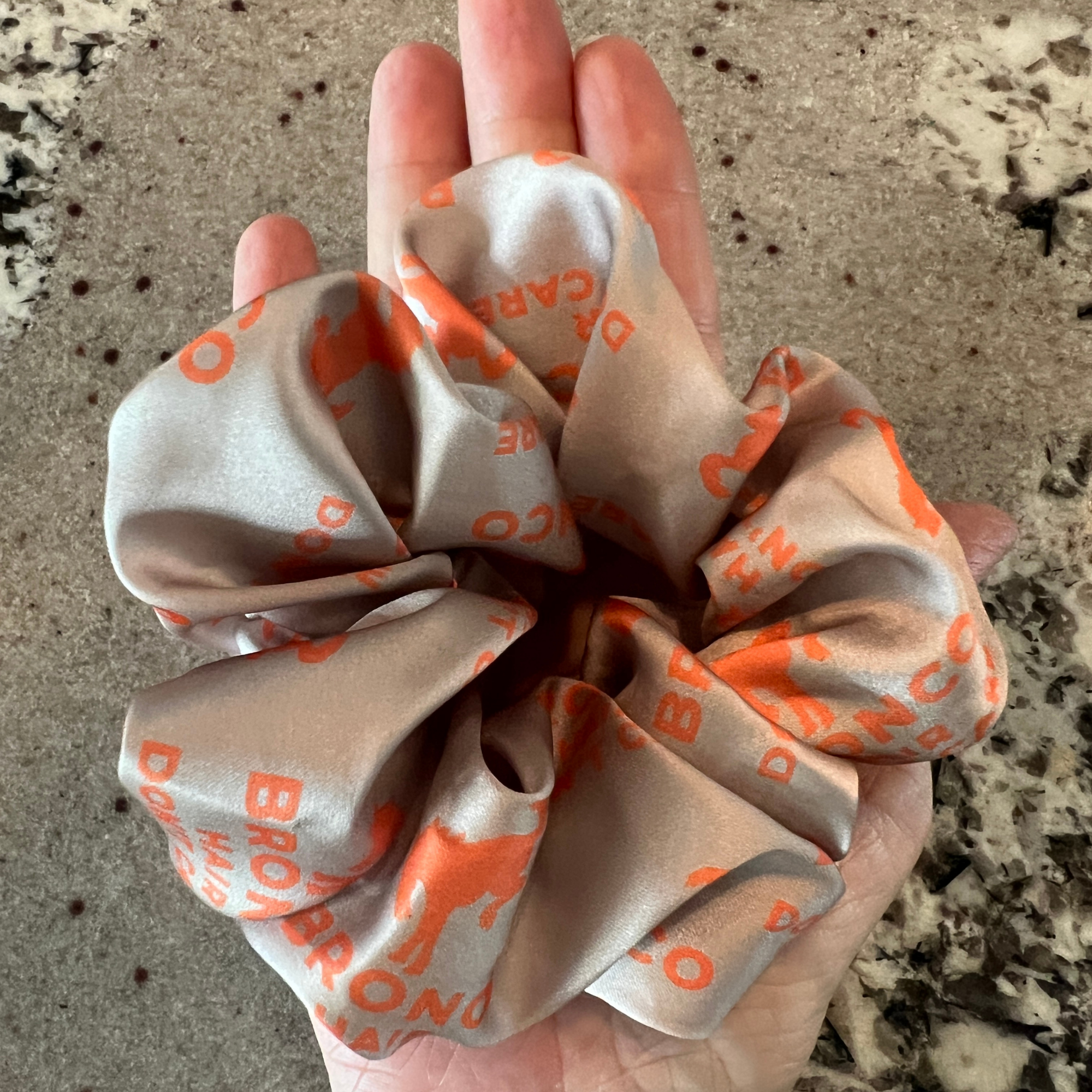 Bronco Hair Don't Care satin scrunchie in tan and coral shown in hand for size reference.