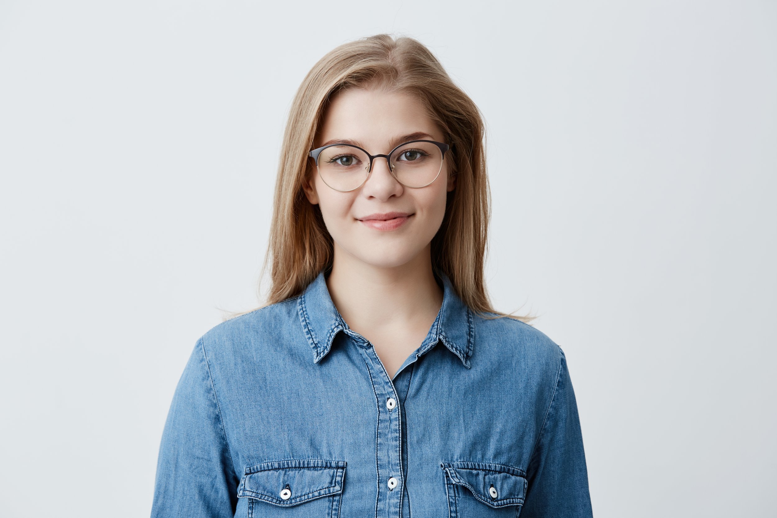A young woman with long blonde hair, wearing glasses and a denim shirt, smiling against a plain light background.
