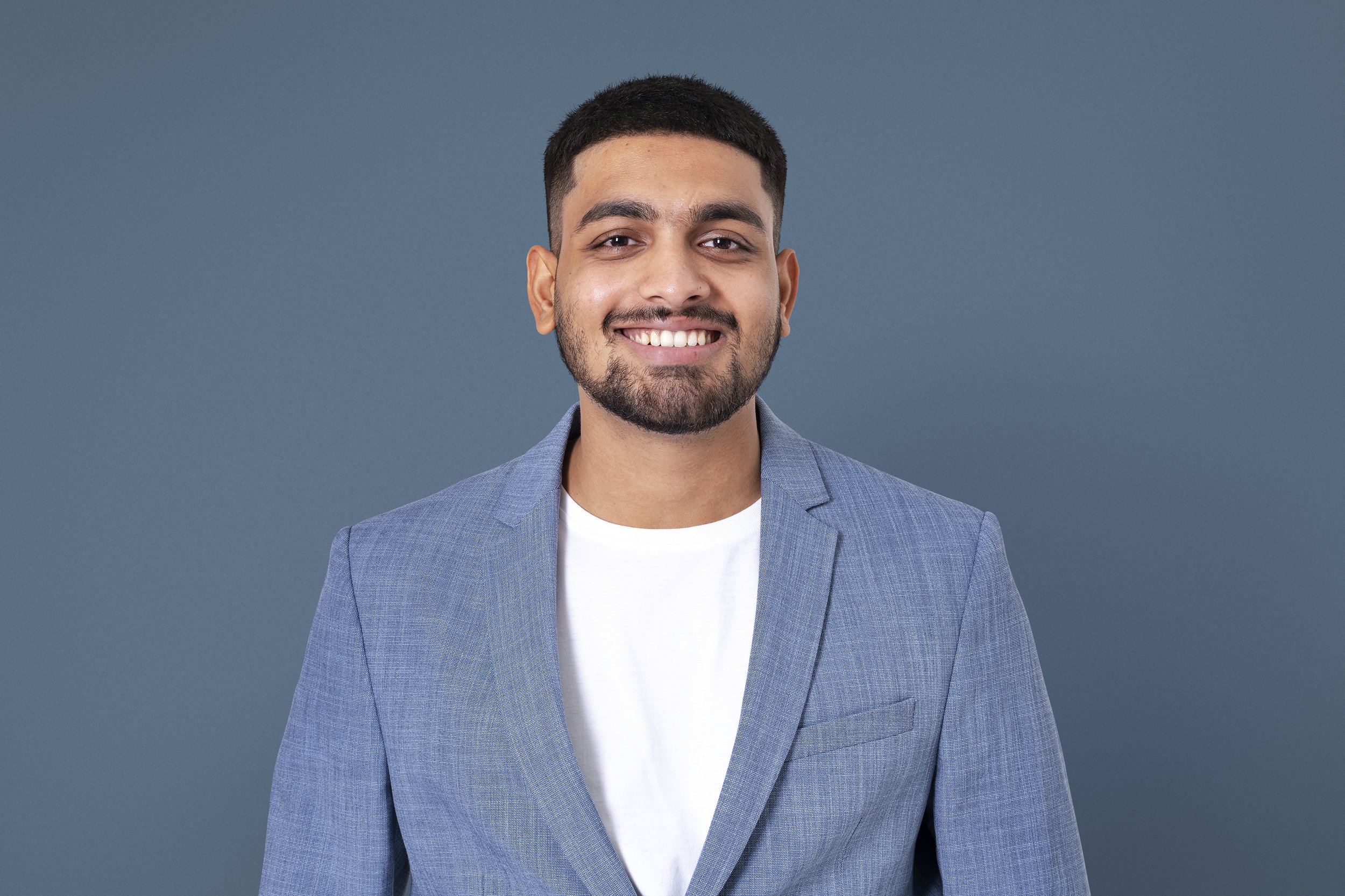 Young man with short dark hair and a beard, smiling, wearing a light blue blazer over a white shirt, standing against a plain blue background.