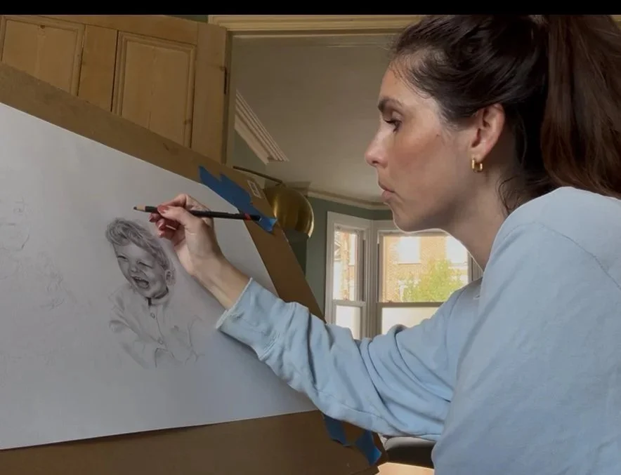A woman drawing a child's portrait on a large sheet of paper in a cozy room with large windows and wooden cabinets.