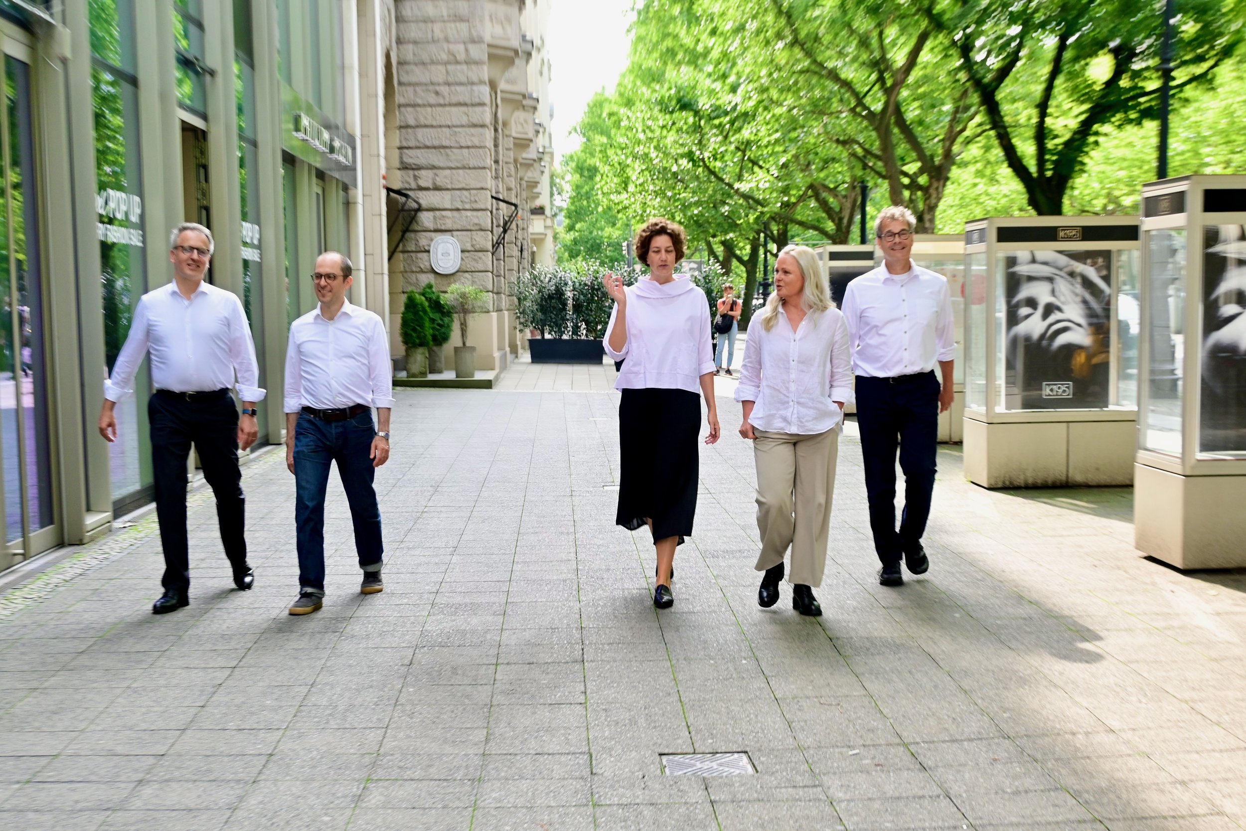 Fünf Menschen, vier Männer und eine Frau, gehen auf einer Stadtstraße zwischen Bäumen und Gebäuden. Sie tragen formelle Kleidung und scheinen sich zu unterhalten.
