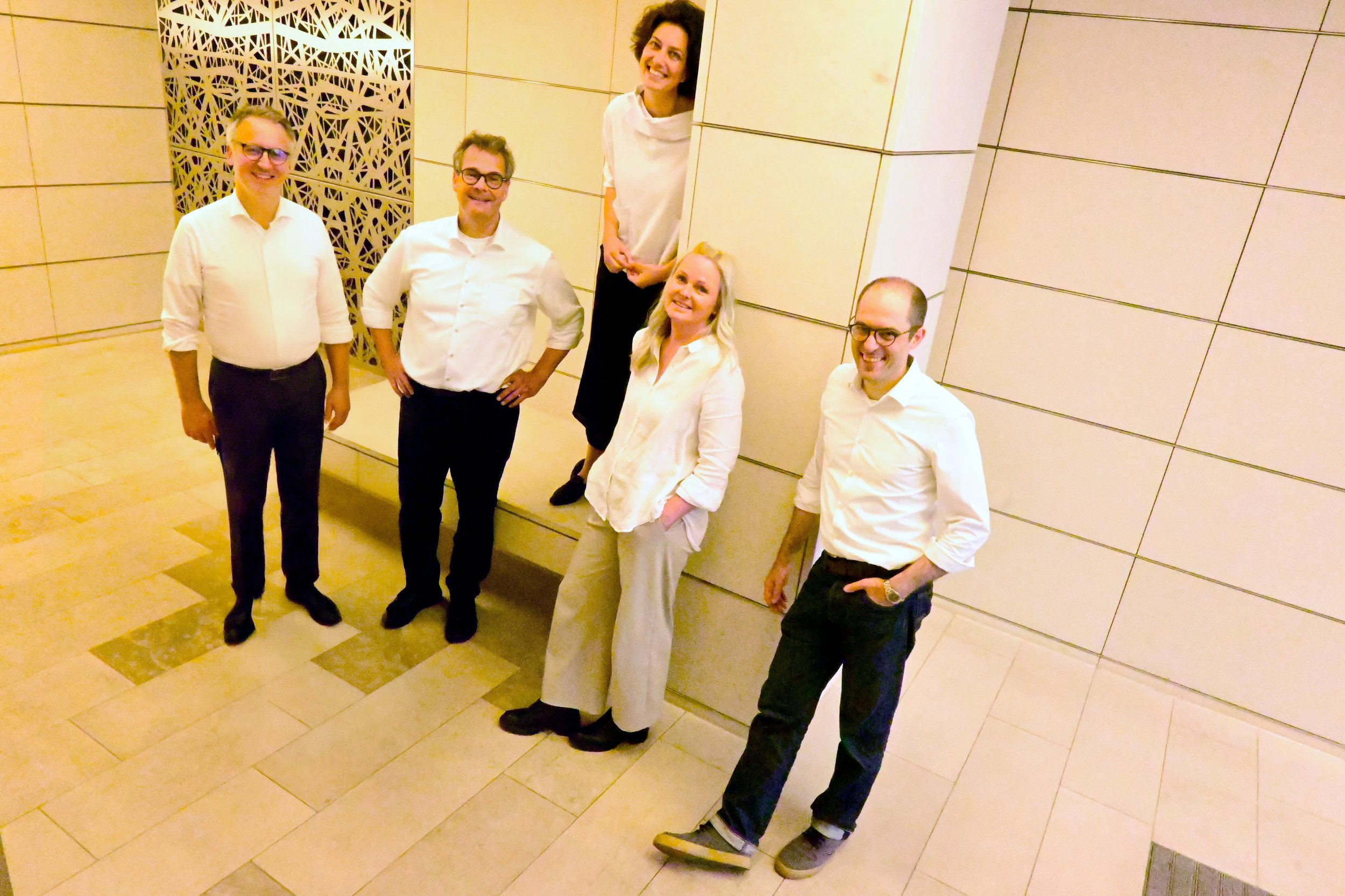 Fünf Menschen, drei Männer und zwei Frauen, posieren in einem Raum mit hellen Wänden und Bodenfliesen, einige von ihnen tragen Brillen und alle sind in weißer oder heller Kleidung, lächeln und scheinen sich in angenehmer Stimmung zu befinden.