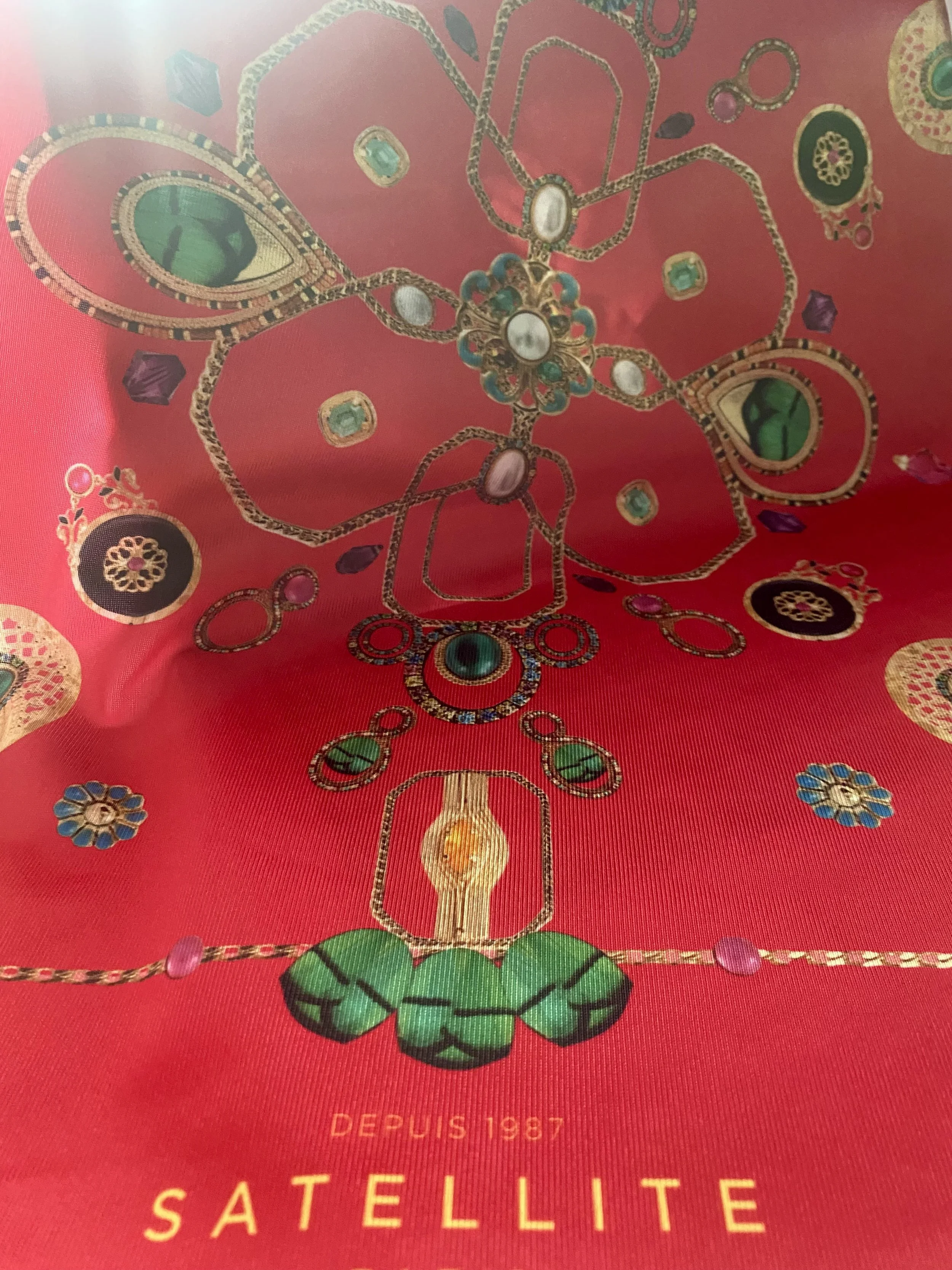 Une image représentant un design de bijoux avec des pierres précieuses sur un fond rouge. En bas, il y a l'inscription "DEPUIS 1987 SATELLITE".