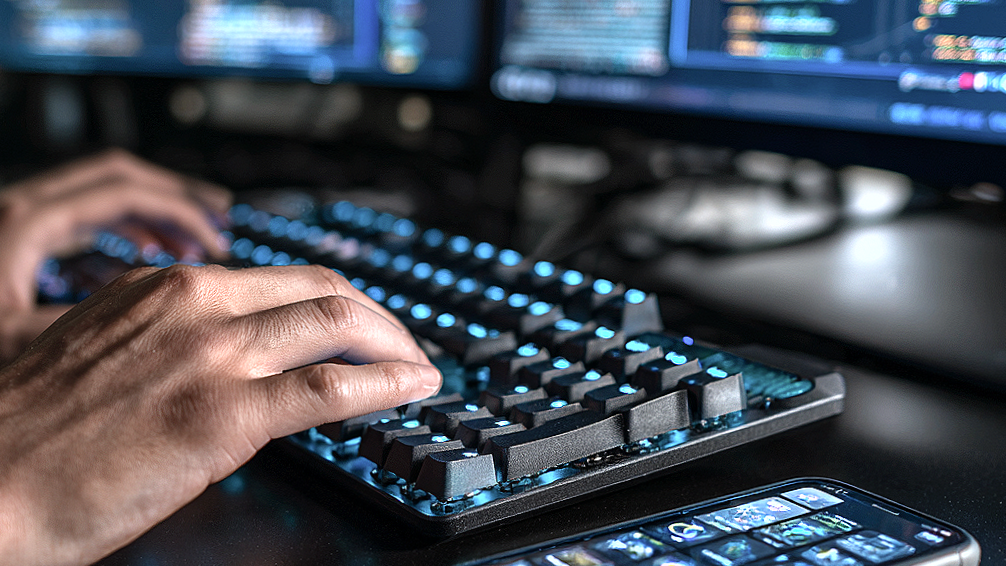 Nahaufnahme der Hände einer Person, die auf einer beleuchteten Computertastatur tippt, mit mehreren Monitoren im Hintergrund.