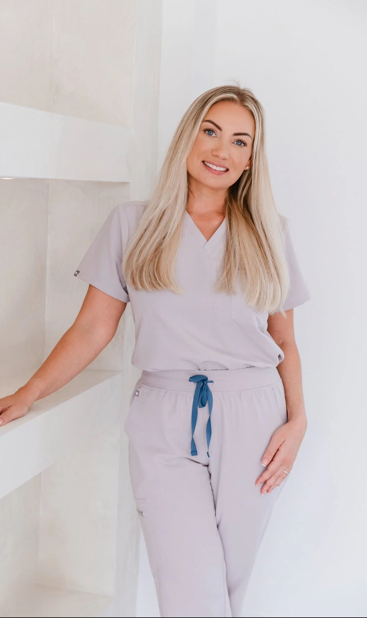 A woman with long blonde hair, wearing light-colored scrubs, smiling and leaning against a white wall.
