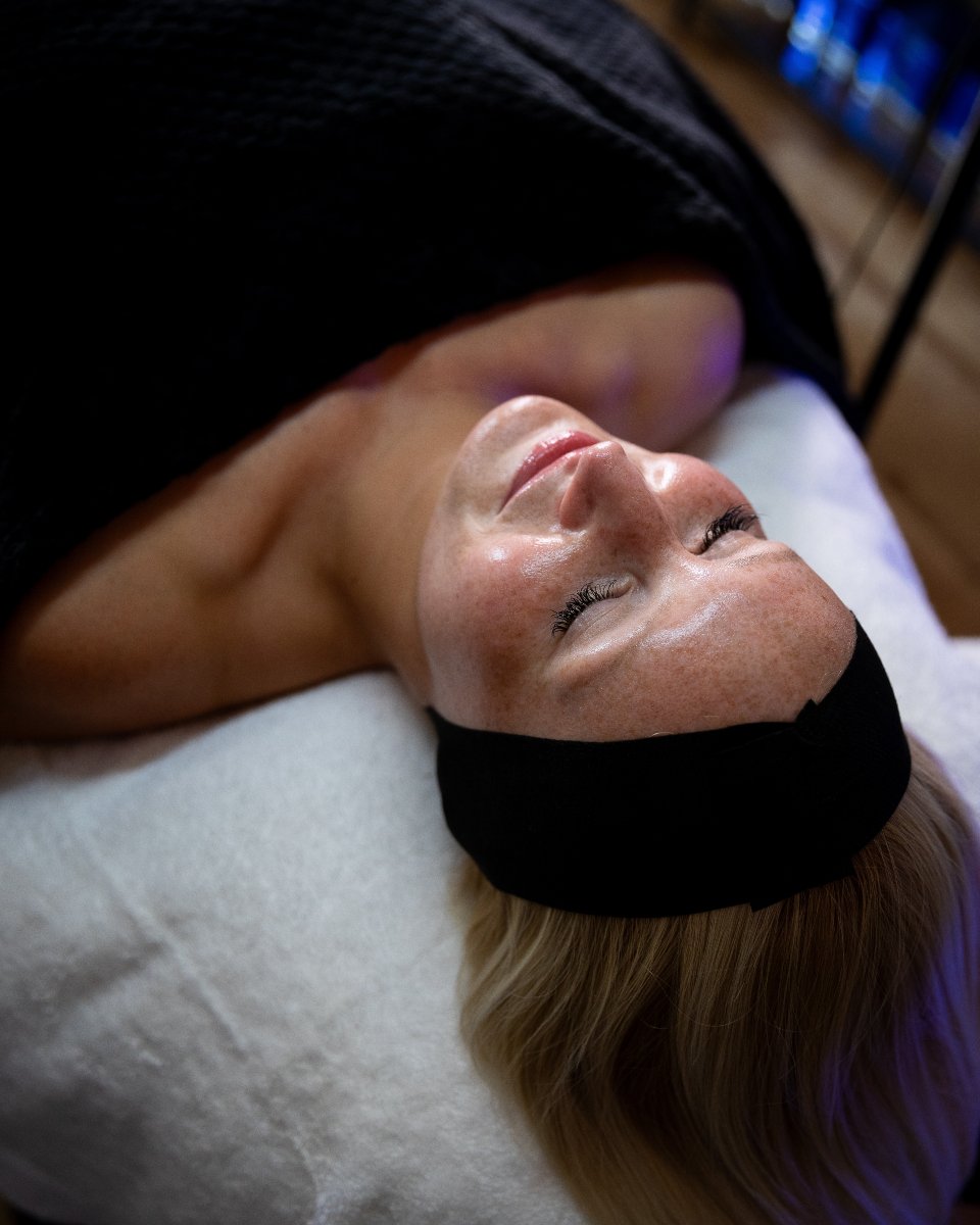 A woman lying on her back on a treatment bed with her eyes closed, wearing a black eye mask, and smiling gently.