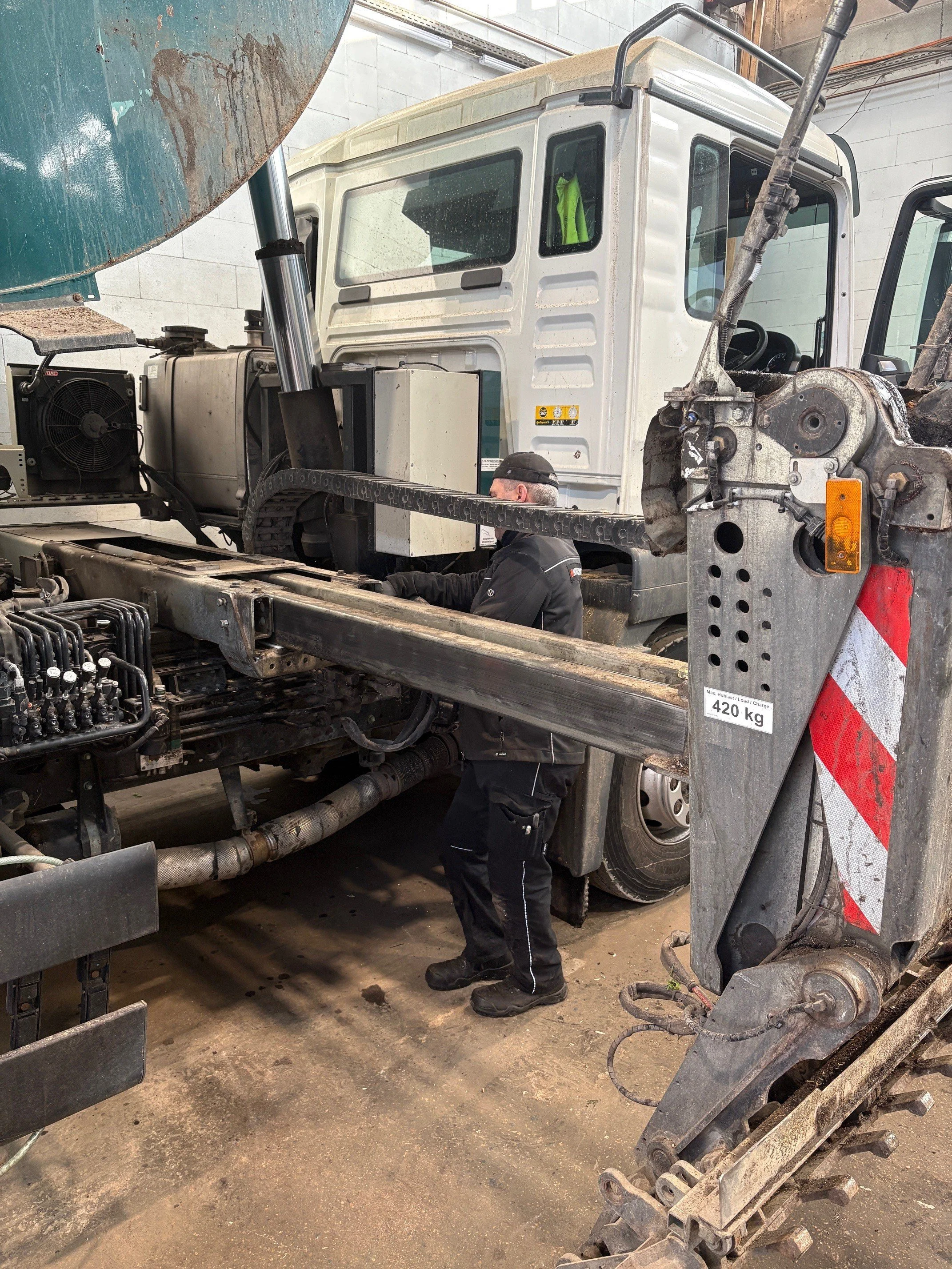 Arbeiter repariert einen großen LKW in einer Werkstatt. Der LKW ist weiß, mit einem Kranarm auf der Seite. Das Fahrzeug wiegt 420 kg.