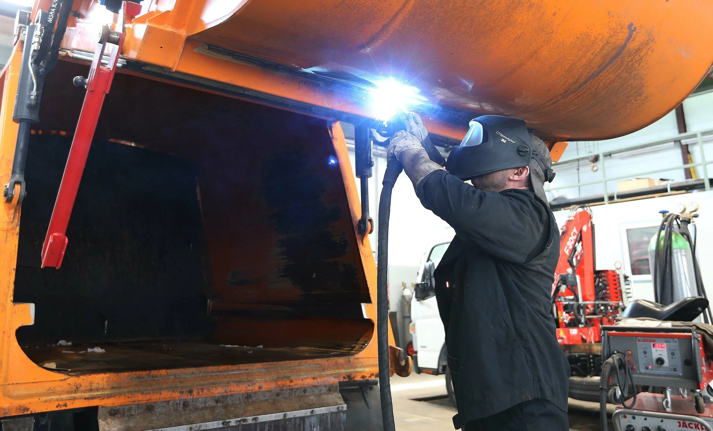 Ein Arbeiter schweißt Metall an einer orangefarbenen Maschine in einer Werkstatt.