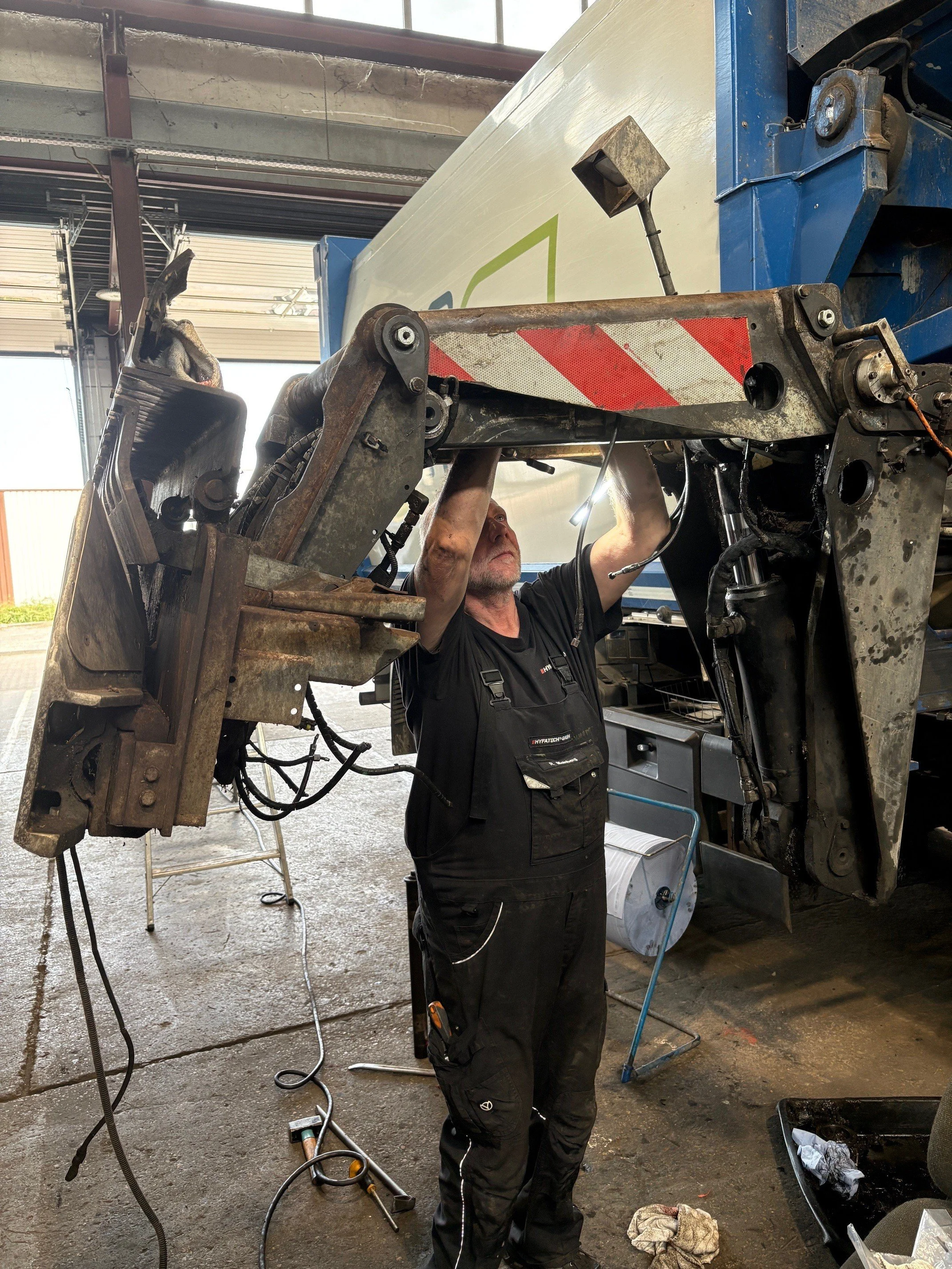 Ein Mechaniker arbeitet an einem großen Fahrzeug, möglicherweise einem LKW, im Werkstattgarage. Er steht auf einer Leiter und führt Wartungsarbeiten oder Reparaturen durch.