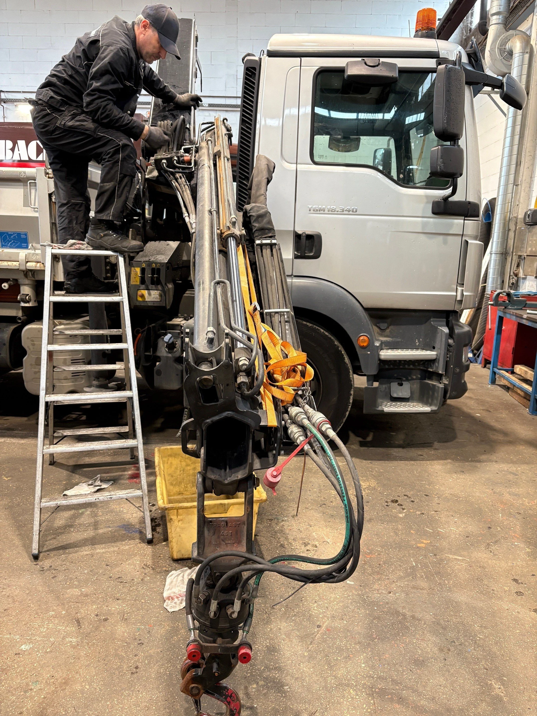 Ein Mann arbeitet auf einem Leiter an einem großen Fahrzeug im Werkstatt. Das Fahrzeug ist vermutlich ein Kran. Der Mann trägt eine schwarze Jacke, Handschuhe und eine Mütze.