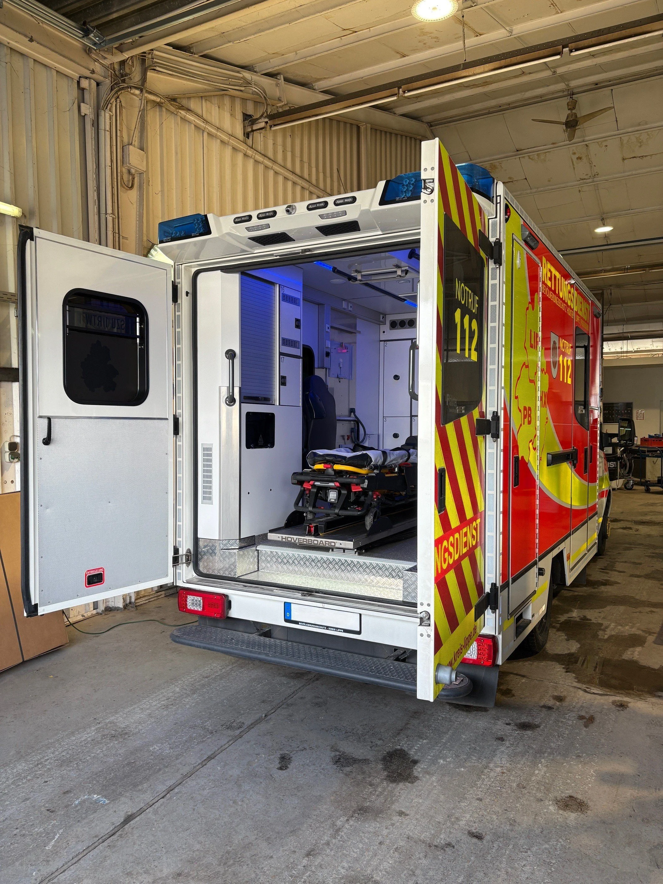 Innenraum eines Rettungswagens mit Krankenliegen, medizinischen Geräten und Schubladen. Außen gelb-rote Warnmarkierungen und die Aufschrift 'Rettungsdienst'.