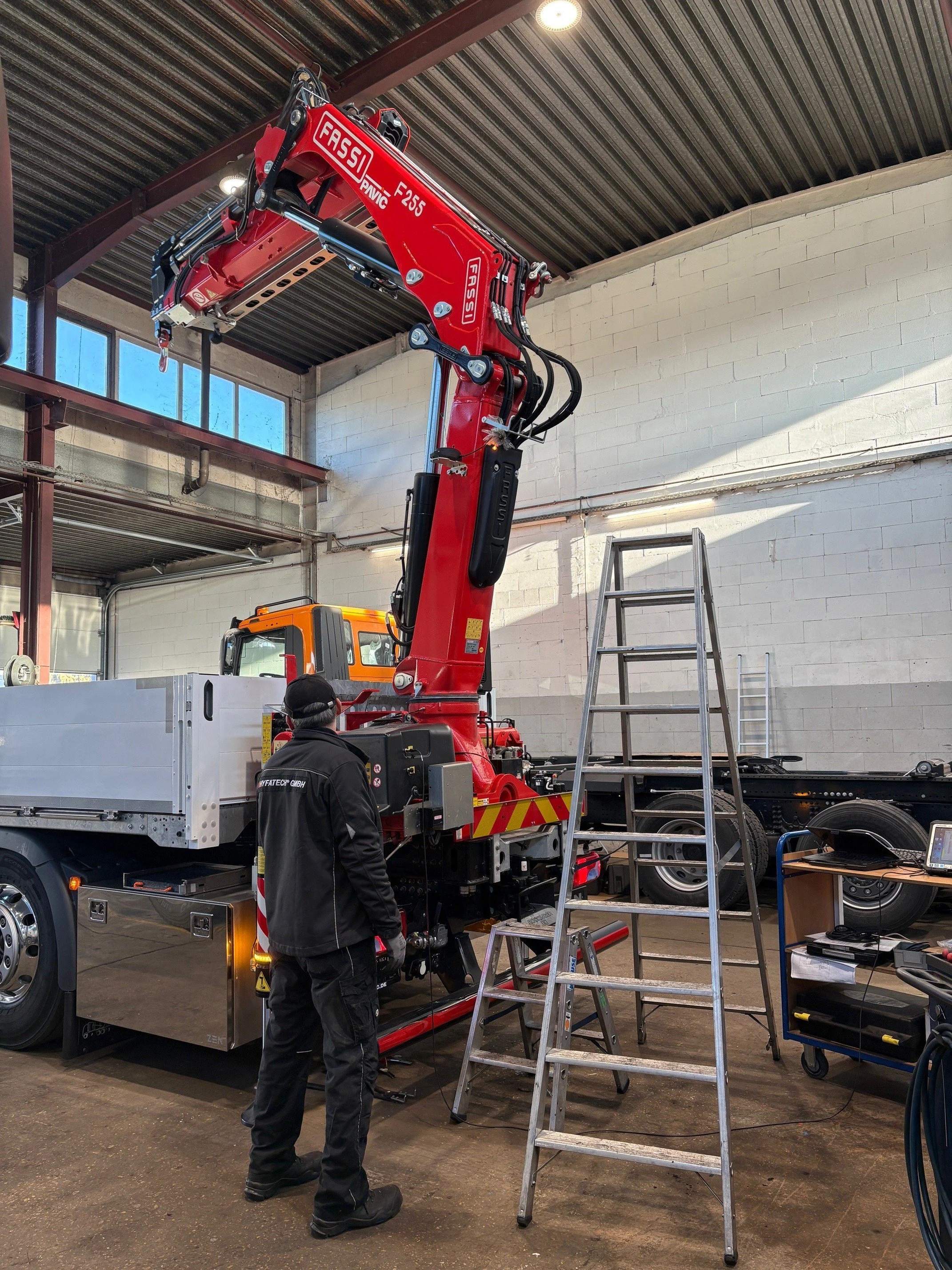 Ein Techniker steht vor einem roten Kran mit der Bezeichnung FASSI F255, der in einer Werkstatt oder einem Fabrikationsraum steht. Der Kran ist auf einem Lieferwagen montiert, und es gibt eine Leiter, die davor steht, sowie Werkzeuge und Geräte in der Nähe.
