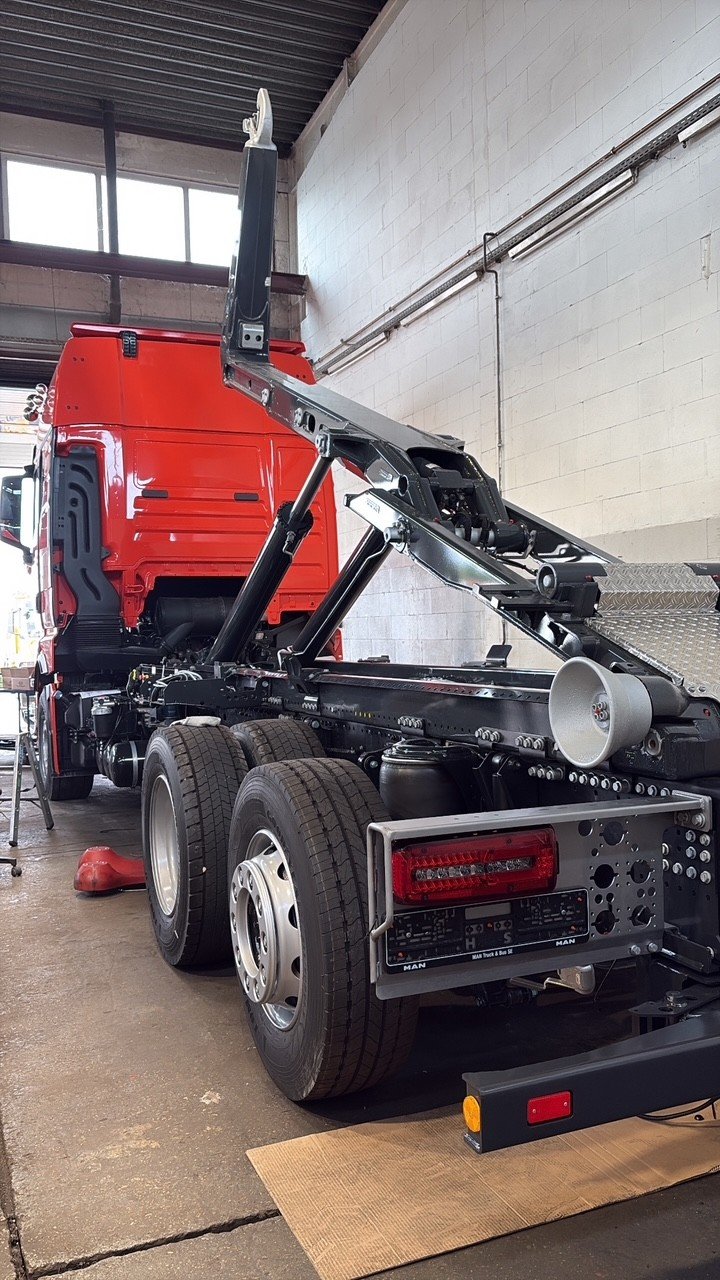 Ein roter LKW im Ausbau in einer Werkstatt, mit sichtbar unmontierten Komponenten und Werkzeugen, wenig Umfeldbeleuchtung, Hochregale und Fenster im Hintergrund.