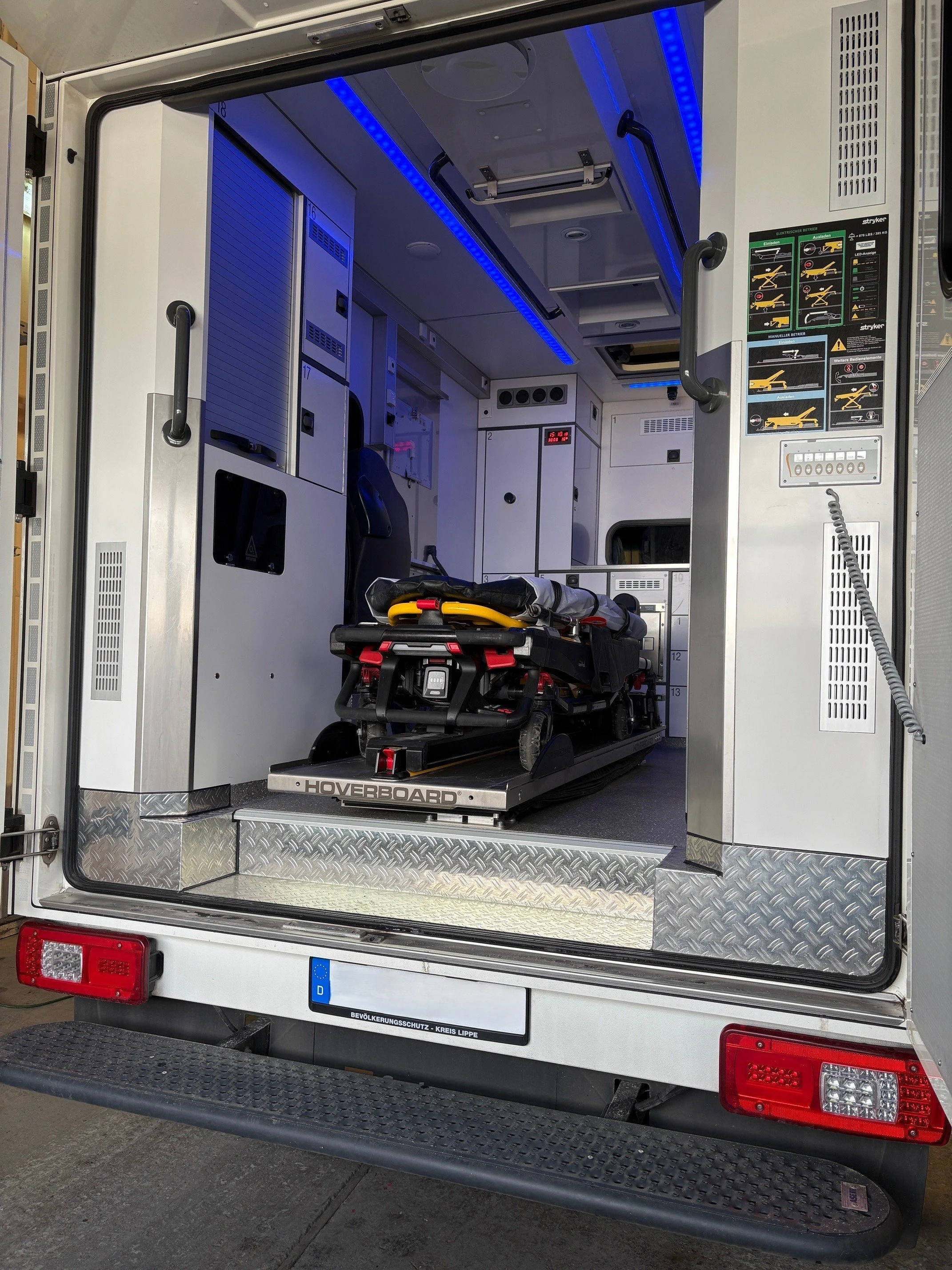 Innenraum eines Rettungswagen mit einer Trage auf einer speziellen Plattform, blauer LED-Beleuchtung an der Decke, medizinische Geräte und Anleitungen an den Wänden.
