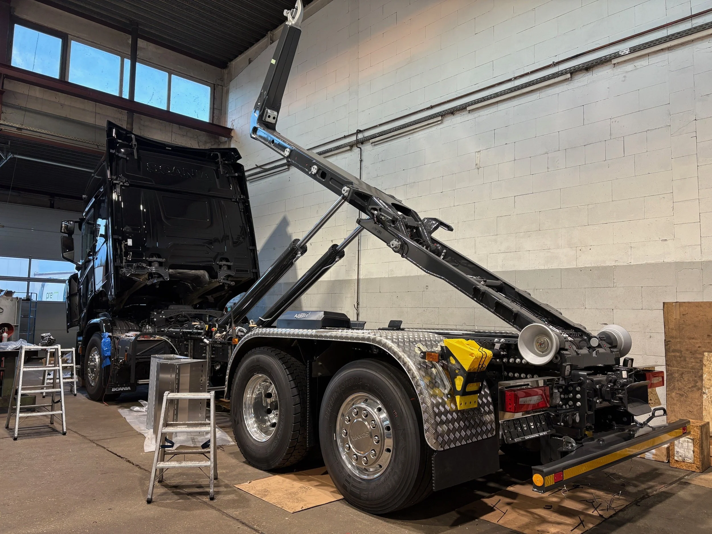 Ein schwarzer LKW in einer Werkstatt, bei dem die Motorhaube geöffnet ist. Der LKW ist aufgebockt, und es sind Werkzeugleitern und Bauteile um das Fahrzeug herum.