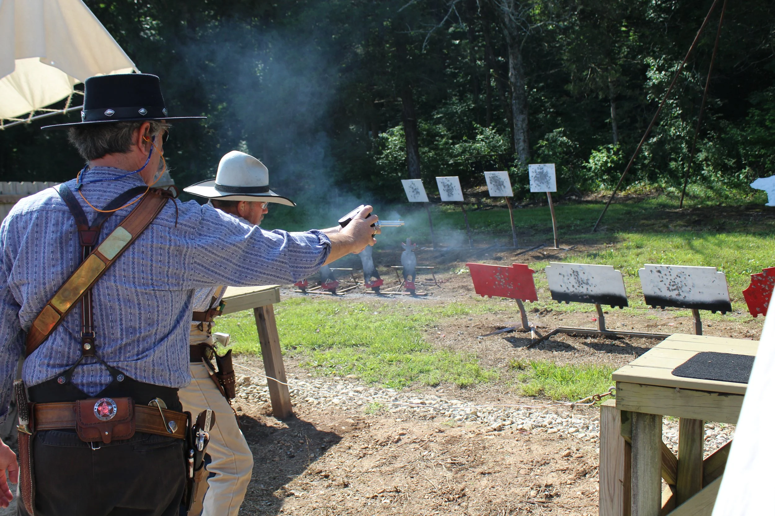 Two men dressed as cowboys, one aiming a pistol at a shooting range with targets set up behind, surrounded by trees and greenery.