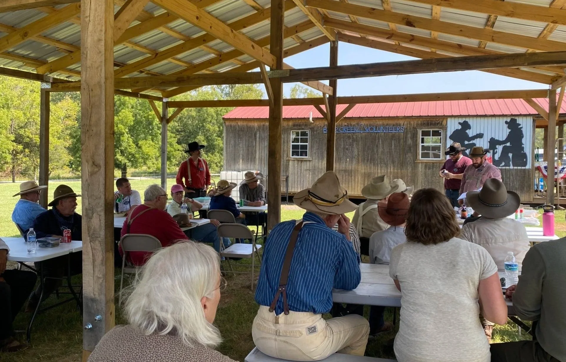 People attending a presentation outdoors under a wooden shelter, seated at tables, wearing wide-brimmed hats, with a wood-paneled building in the background featuring a silhouette mural of two musicians.