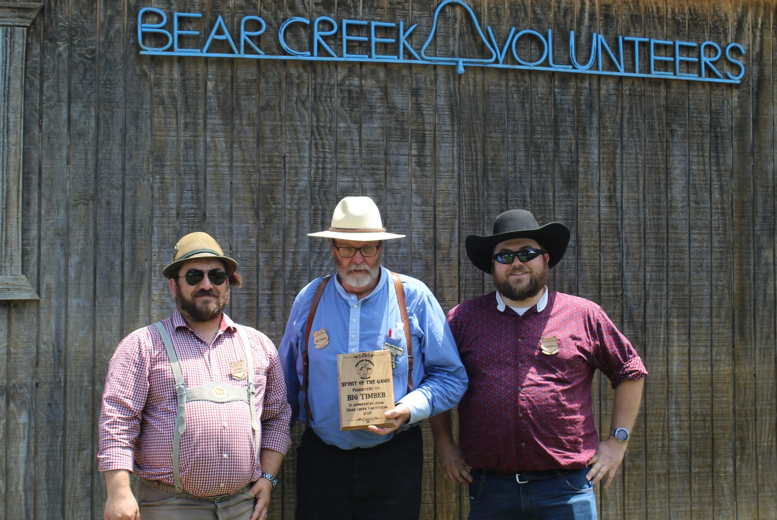 Three men standing outdoors in front of a rustic wooden wall with a neon sign that reads 'Bear Creek Volunteers'. The man in the middle is holding a wooden plaque and is dressed in a blue shirt, suspenders, and a wide-brimmed hat. The man on the left is wearing a checkered shirt, suspenders, sunglasses, and a small hat. The man on the right is wearing a maroon shirt, a large black hat, and sunglasses. All three men are smiling.